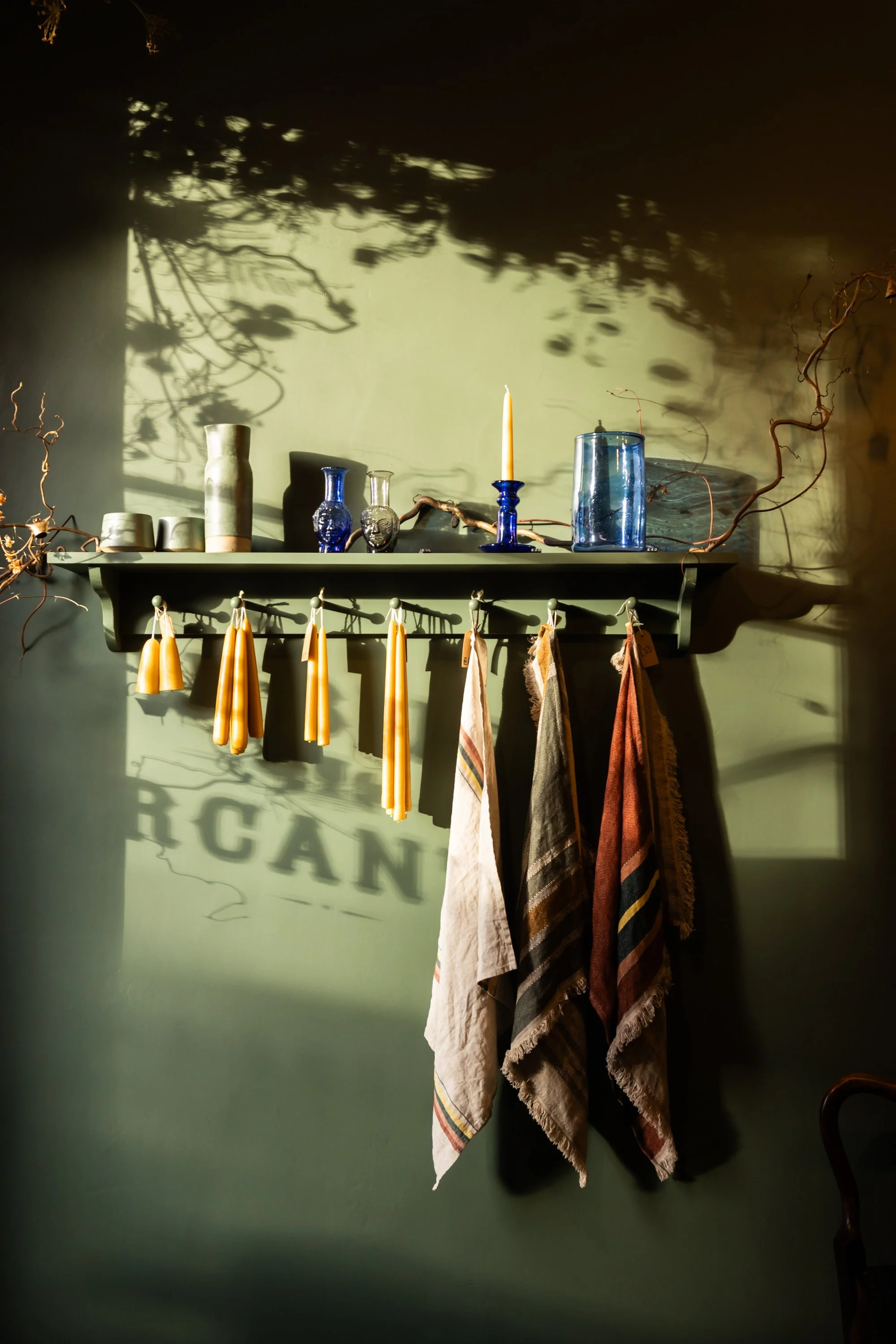 Decorative wall shelf with glass vases, a candle, and hanging towels casting shadows on a green wall.