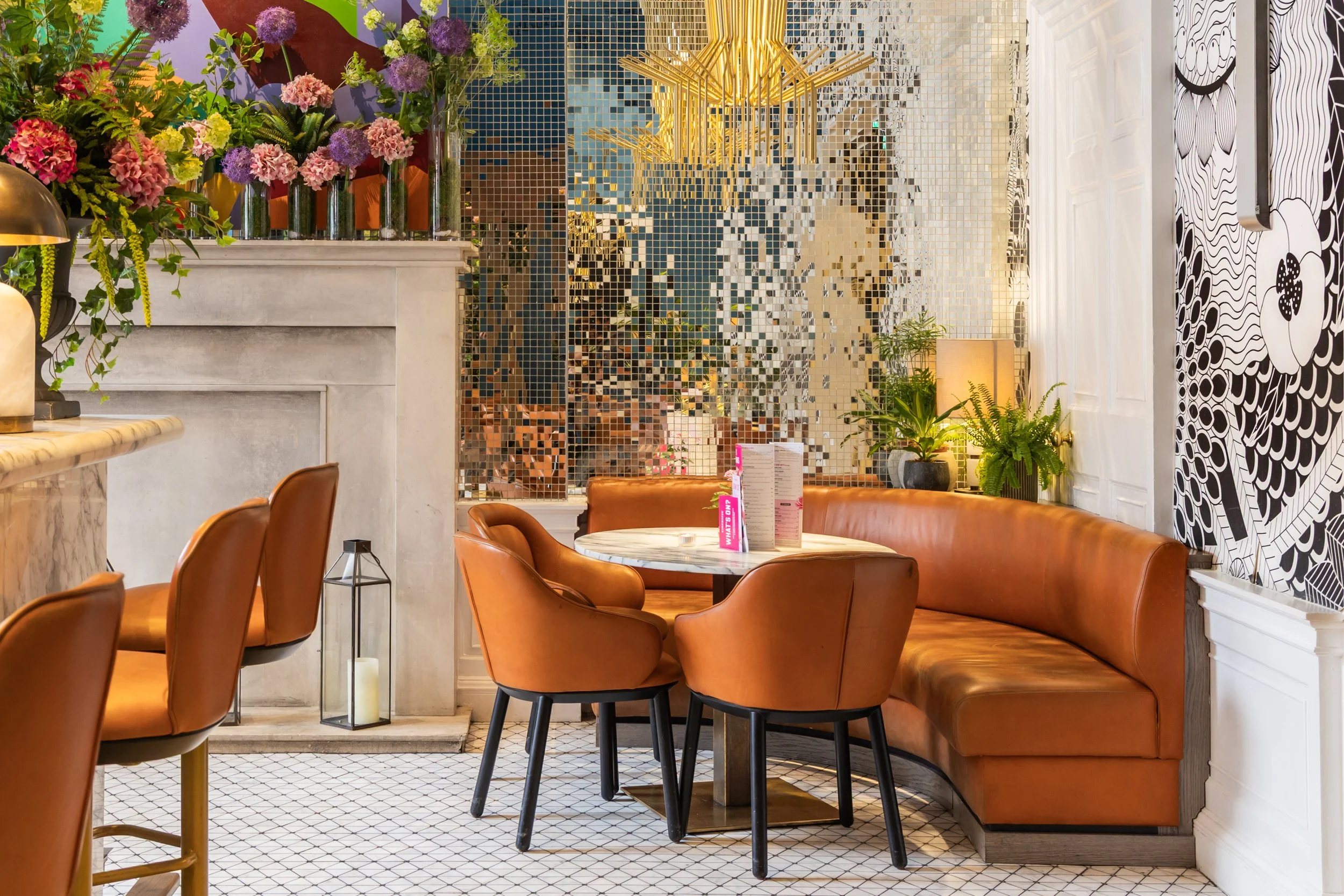 A cozy cafe corner with a white marble fireplace, floral arrangements, a mosaic mirror, and a seating area with a curved leather banquette and round table.