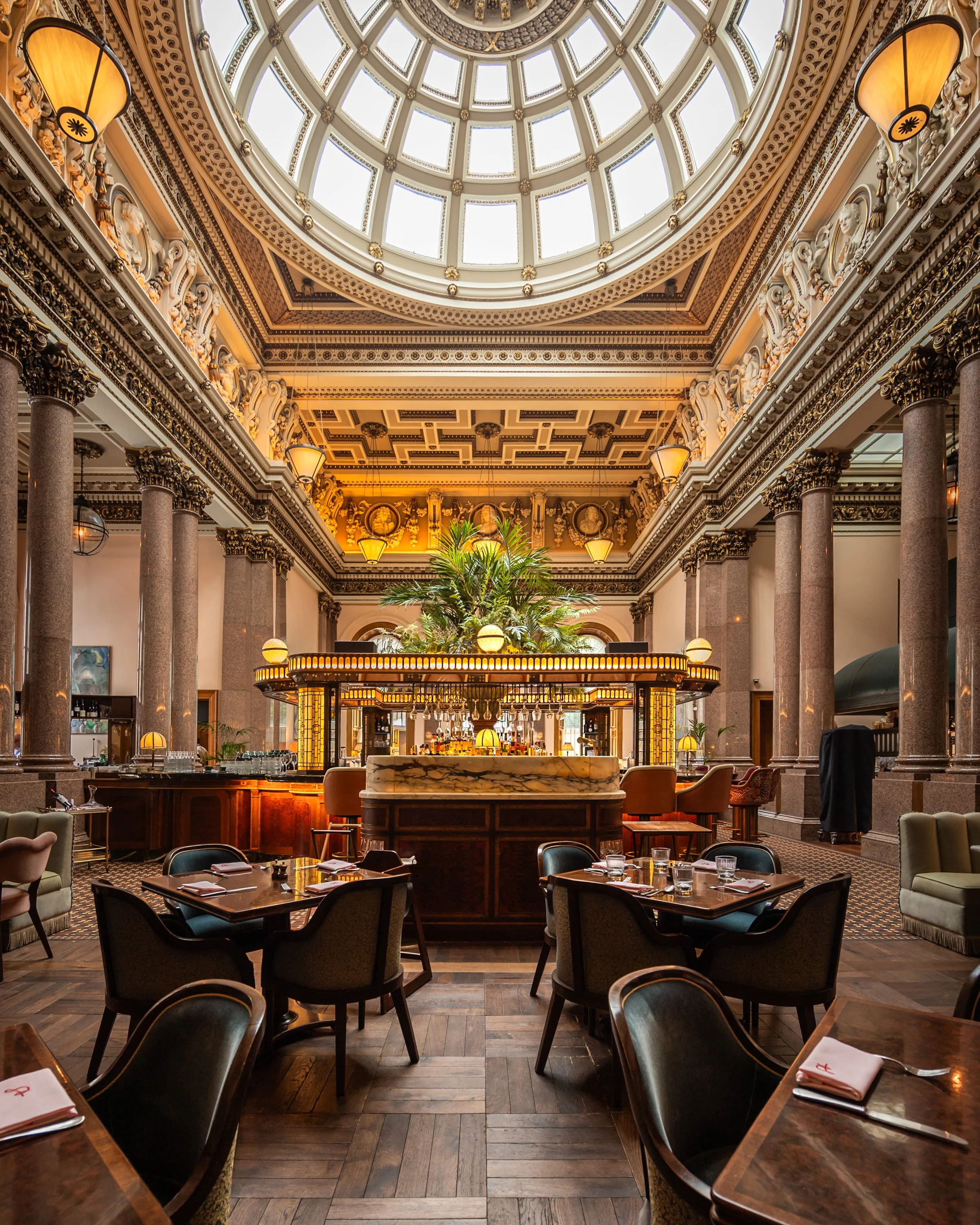 Elegant restaurant with a high domed glass ceiling, ornate architecture, and a central bar area with lush greenery.