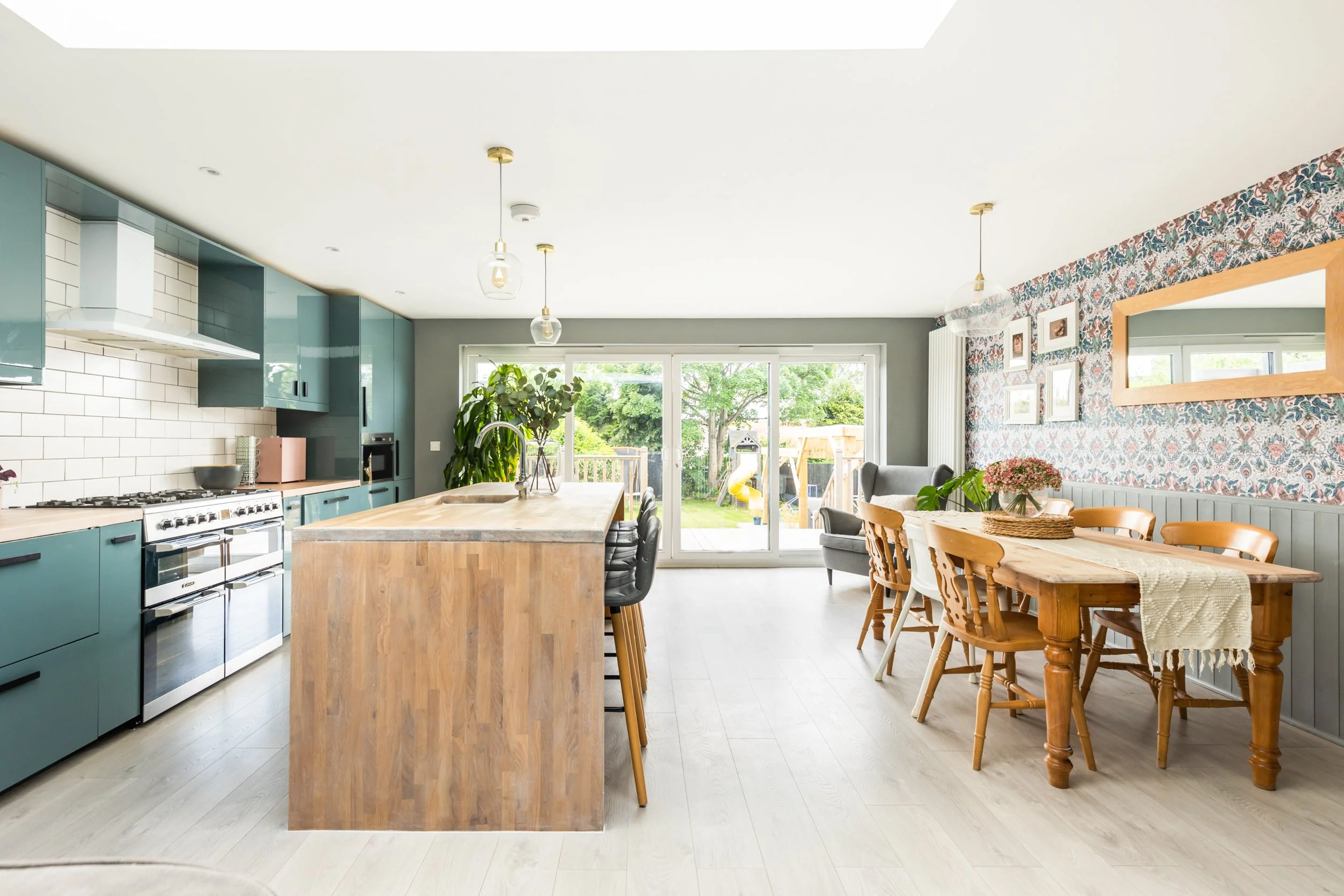 Open-concept kitchen and dining area with large sliding glass doors, teal cabinetry, a wooden island, patterned wallpaper, and a wooden dining table with chairs.