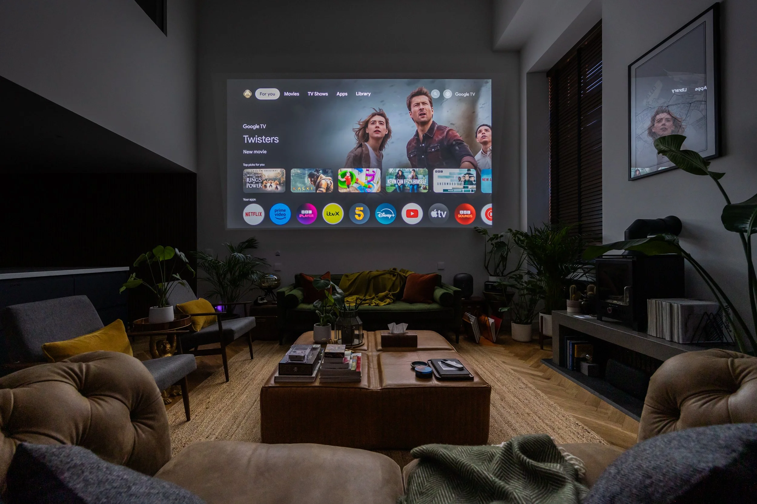 Living room with a large projector screen showing a streaming app interface, surrounded by sofas, chairs, plants, and a coffee table.