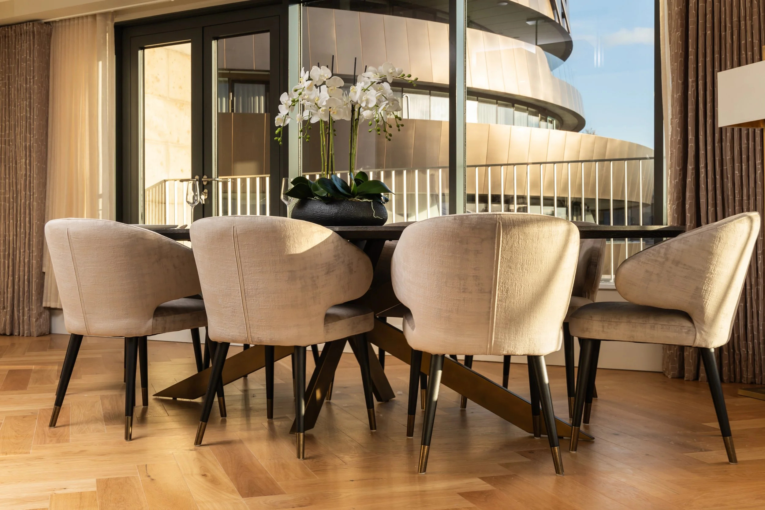A dining room with a round wooden table, six beige upholstered chairs with black and gold legs, and a vase with white orchids on the table. Large windows and a modern building are visible outside.