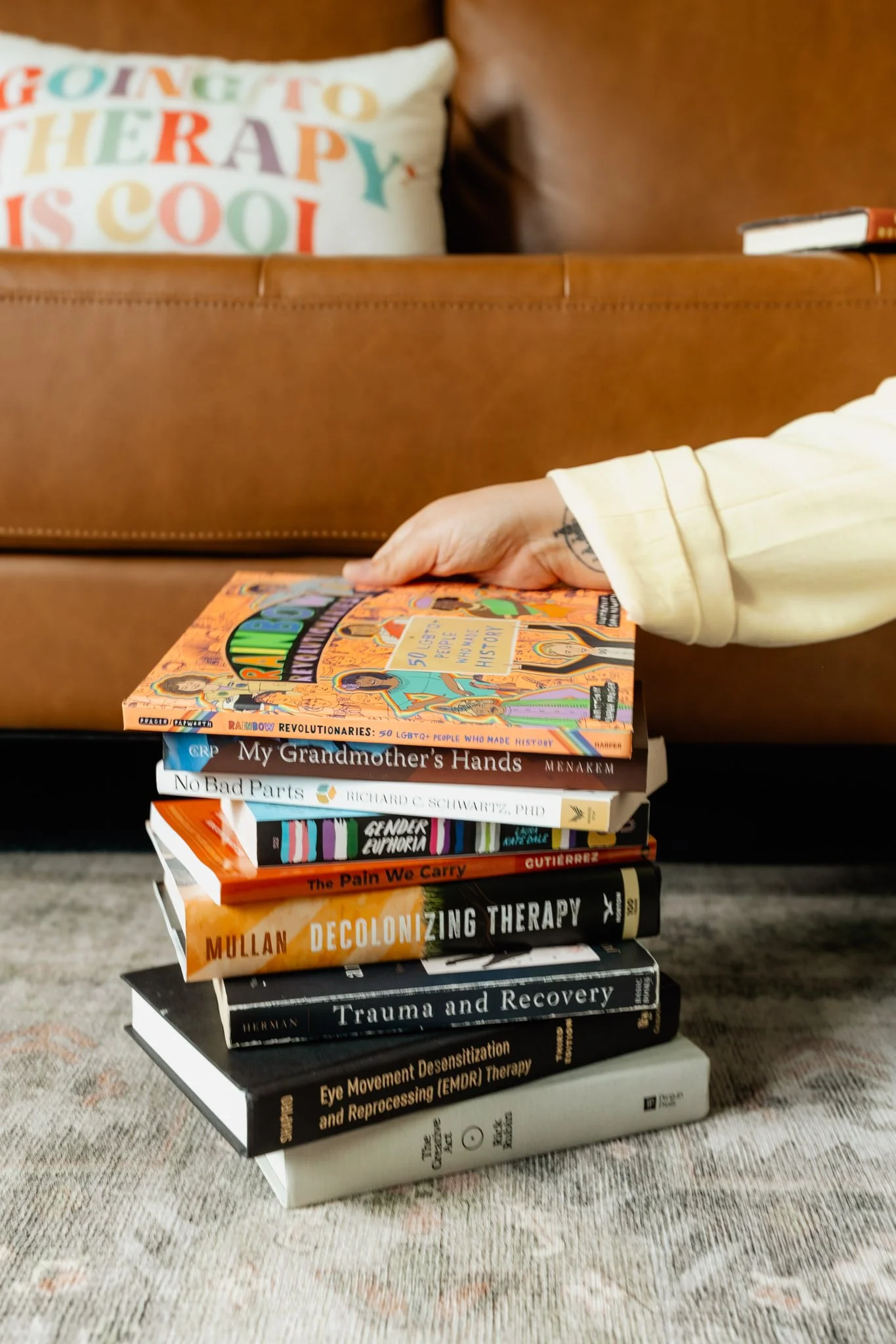 Stack of books on a carpeted floor, with a person's hand reaching for the top book. A brown leather couch with a colorful decorative pillow and a closed book are in the background.