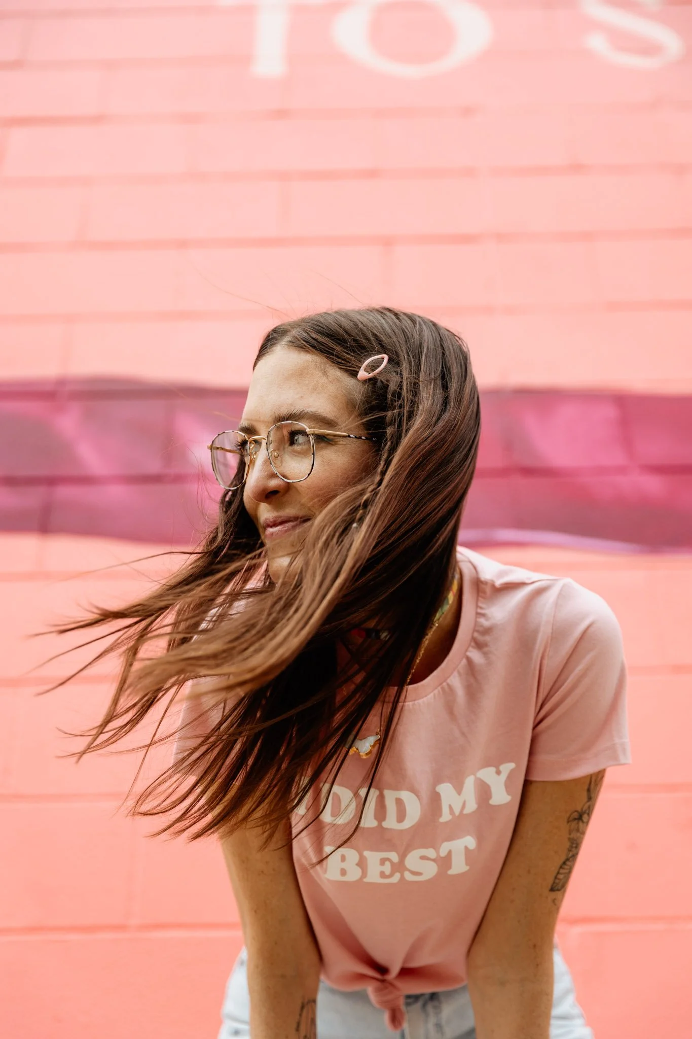 Woman with long brown hair, wearing glasses and a pink hair clip, in front of a pink wall, wearing a pink T-shirt with white text.