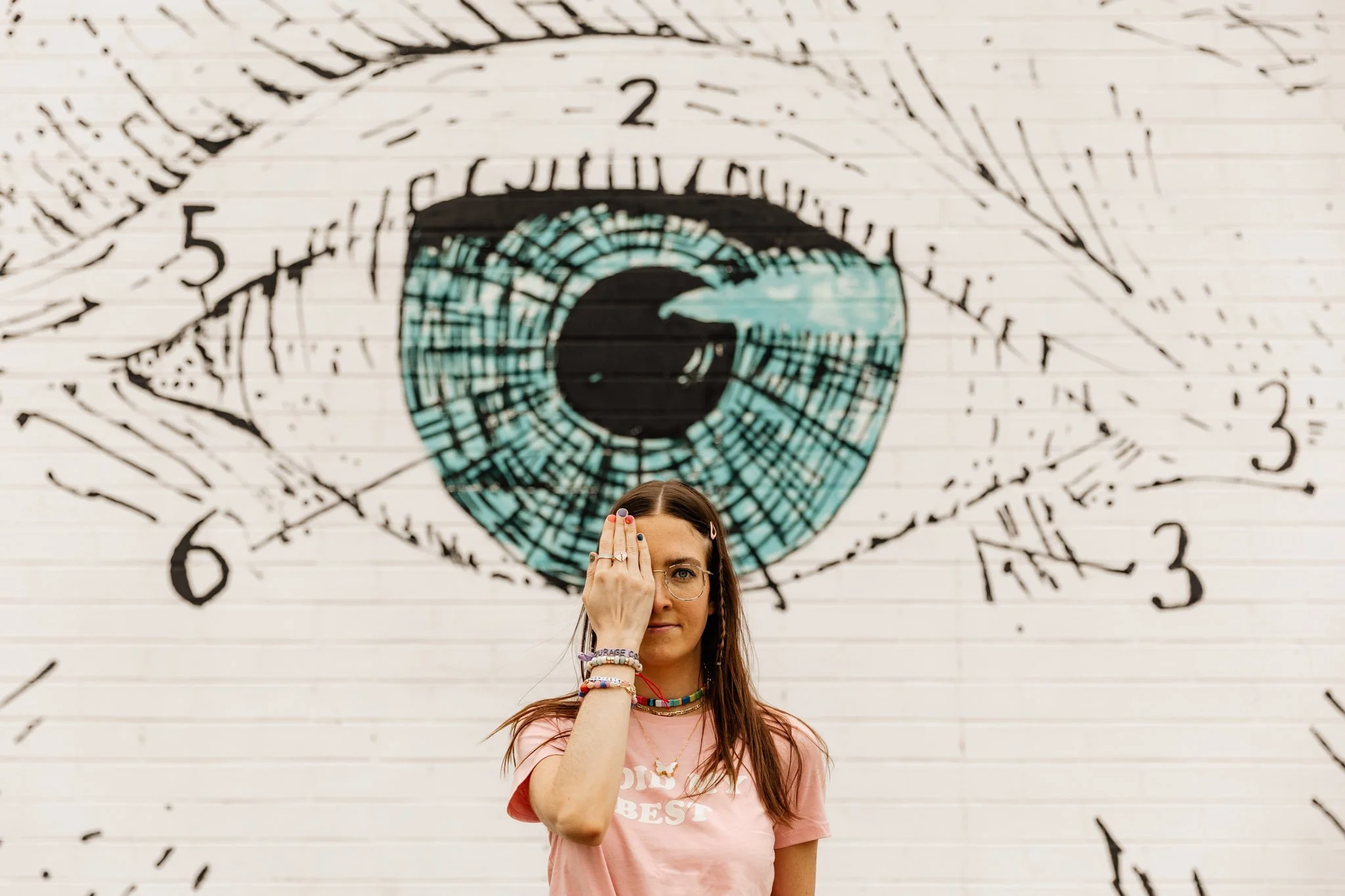 A woman with long hair wearing glasses and multiple colorful bracelets covers one eye with her hand while standing in front of a mural featuring a large eye and handwritten numbers and symbols on a white wall.
