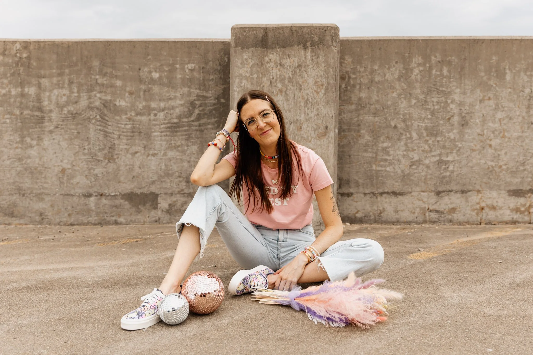 A young woman with glasses, wearing a pink t-shirt, white ripped jeans, and colorful sneakers, sitting on the ground in front of a large concrete wall. She is smiling and resting her head on her hand. There are disco balls, a colorful fluffy rug, and
