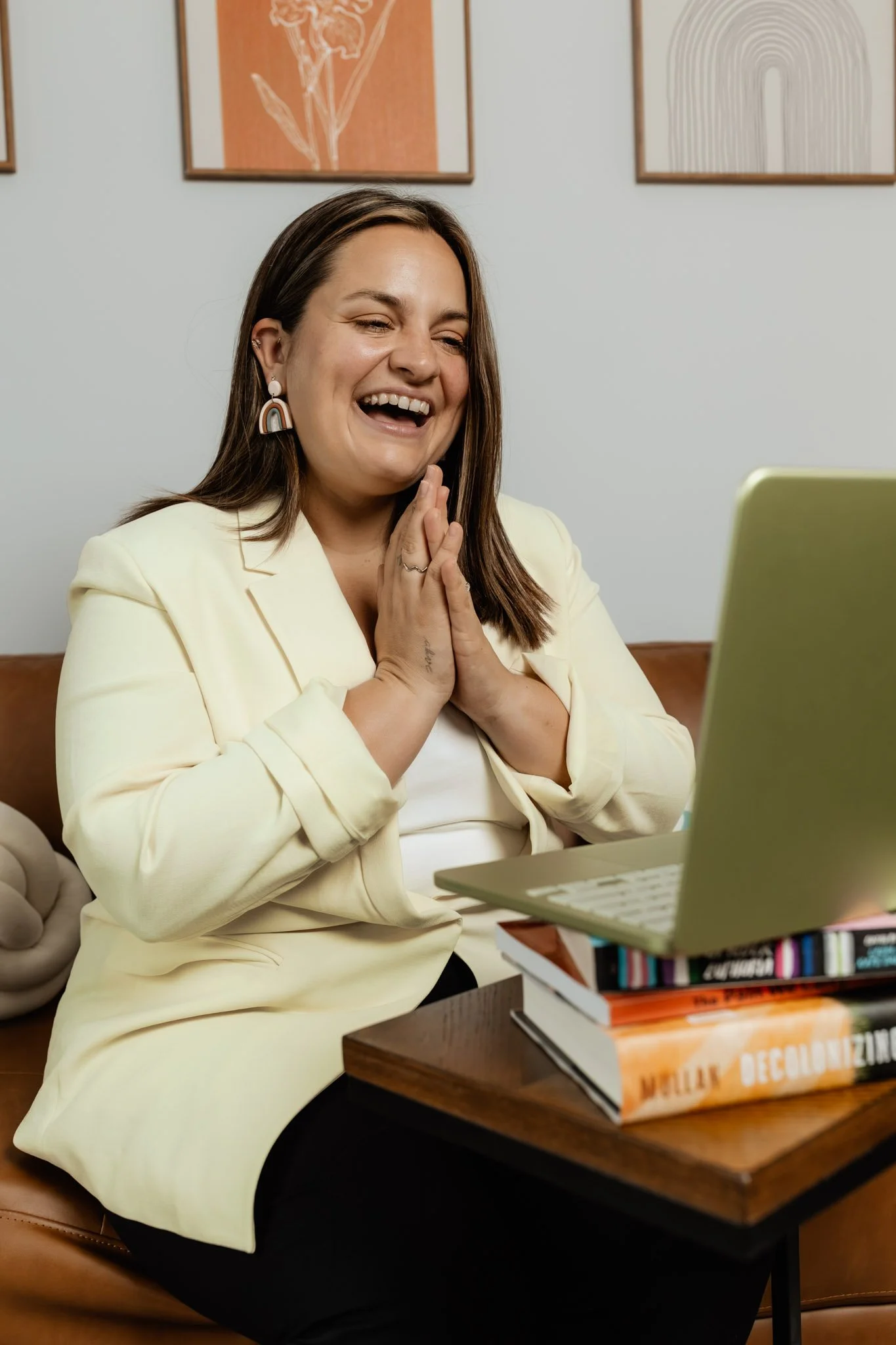A woman with brown hair, wearing a cream blazer and rainbow-shaped earrings, is smiling and looking at a laptop. She has her hands clasped together near her face. She is sitting by a desk with a stack of books and a lemon-colored laptop.