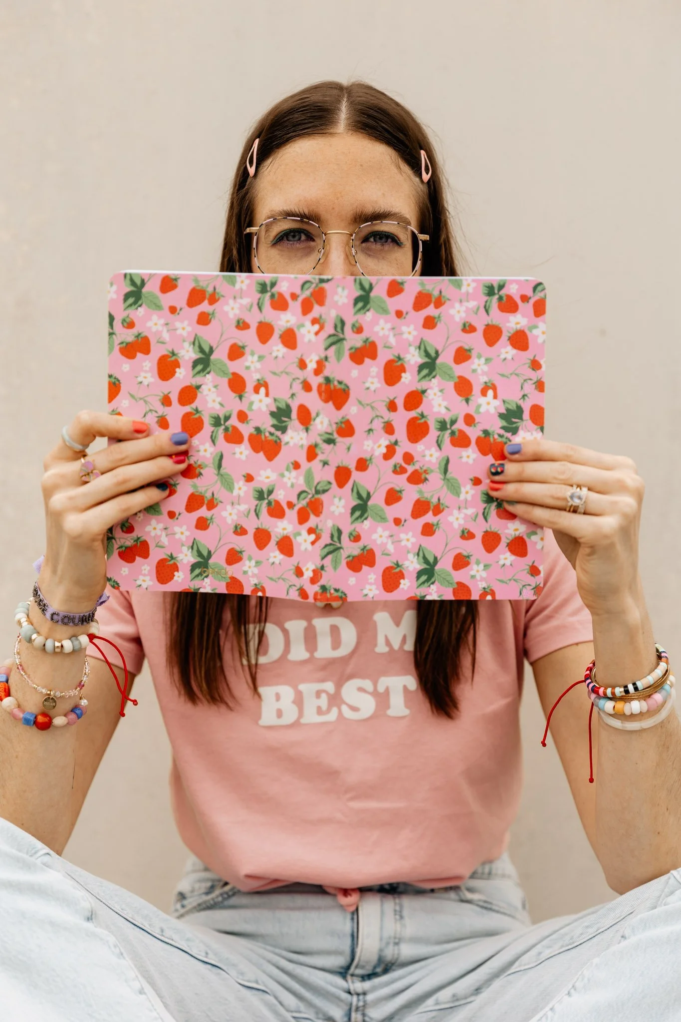 Person with long brown hair, wearing glasses, holding a pink notebook with a strawberry and floral pattern in front of their face. Wearing a pink T-shirt with white text and colorful bracelets on both wrists. Sitting cross-legged, beige wall backgrou