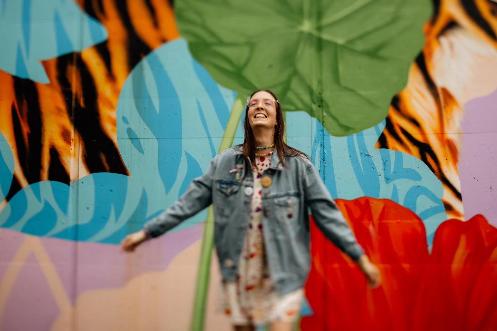 A young woman with glasses smiling and posing in front of a colorful mural featuring large leaves and tiger stripes. She is wearing a denim jacket, a patterned dress, and a beaded necklace.