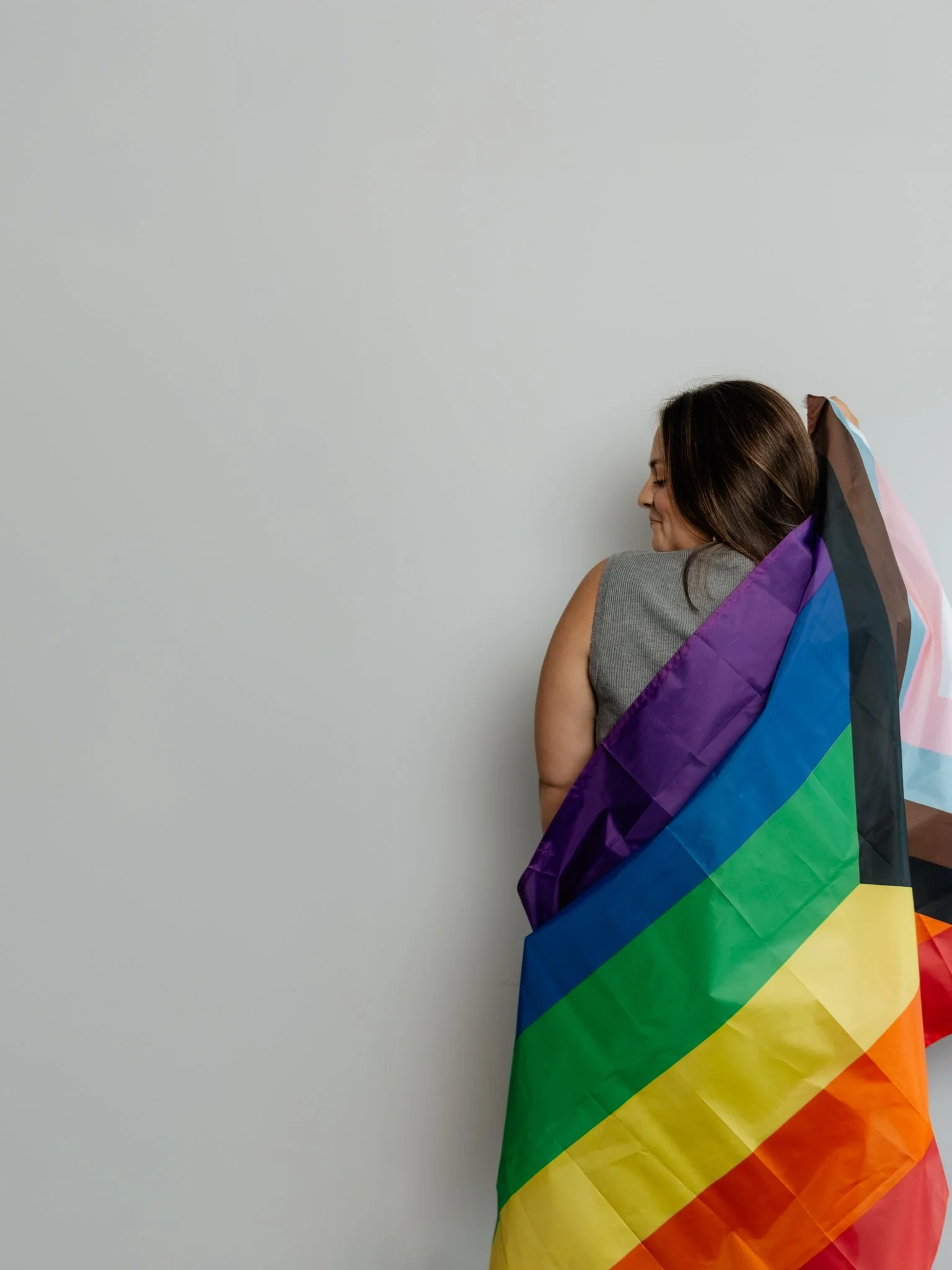 A woman standing against a plain light-colored wall, with a rainbow pride flag draped over her shoulders.