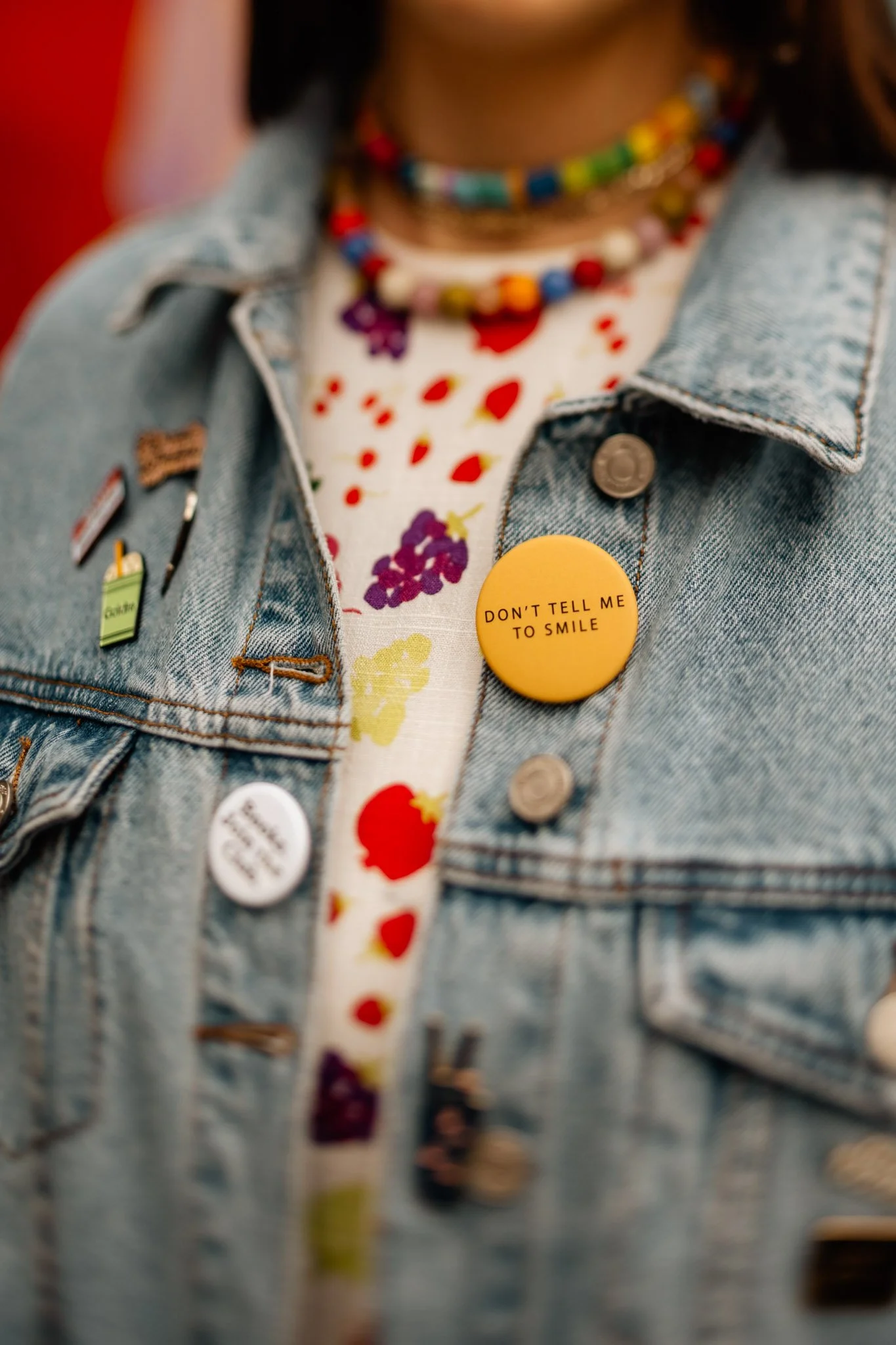 Close-up of a person wearing a denim jacket with several pins, a colorful beaded necklace, and a button that says 'DON'T TELL ME TO SMILE' over a floral-patterned shirt.