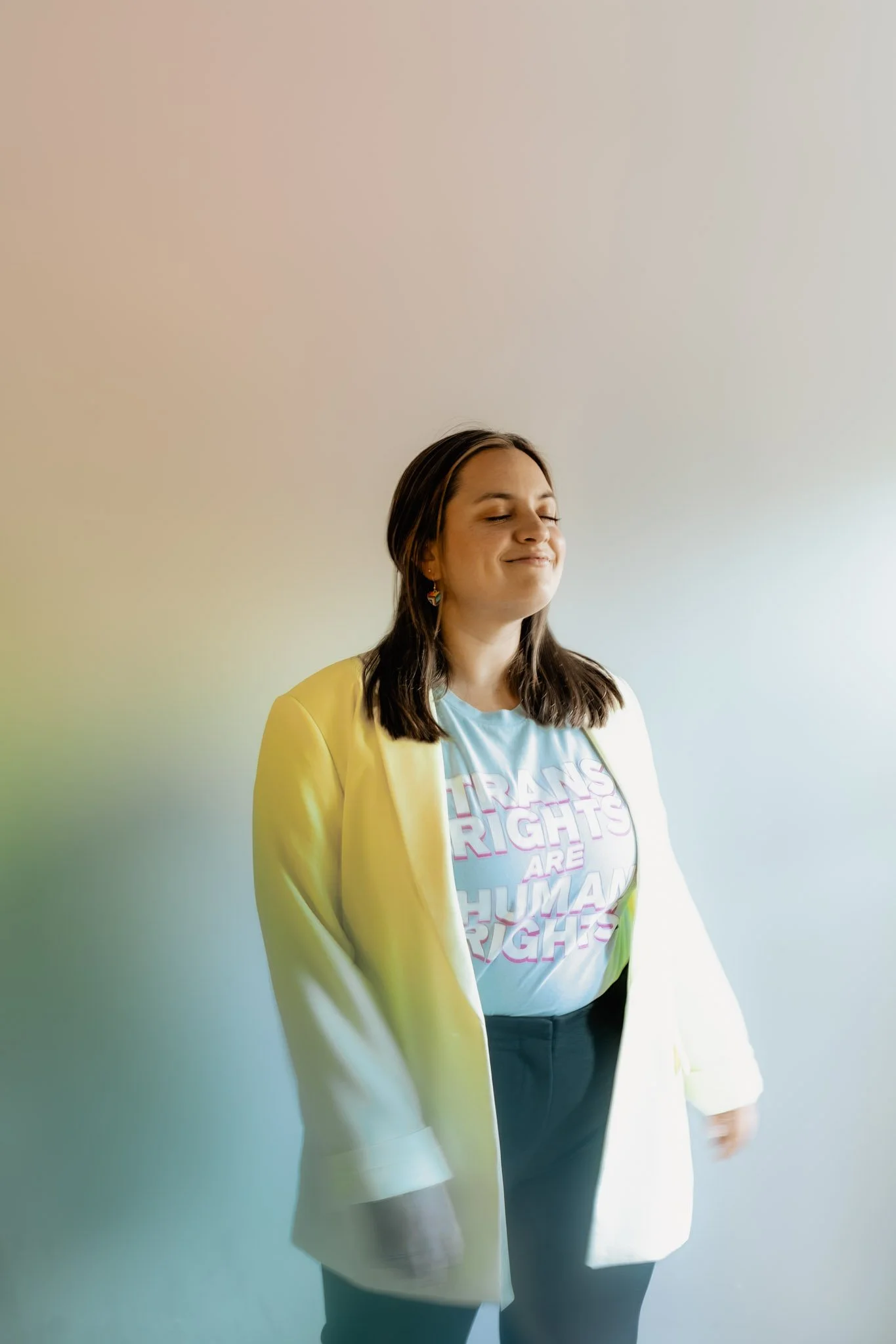 Young woman with long brown hair smiling, standing against a plain gradient background. She is wearing a light blue T-shirt with a colorful graphic and text, and a light-colored blazer.