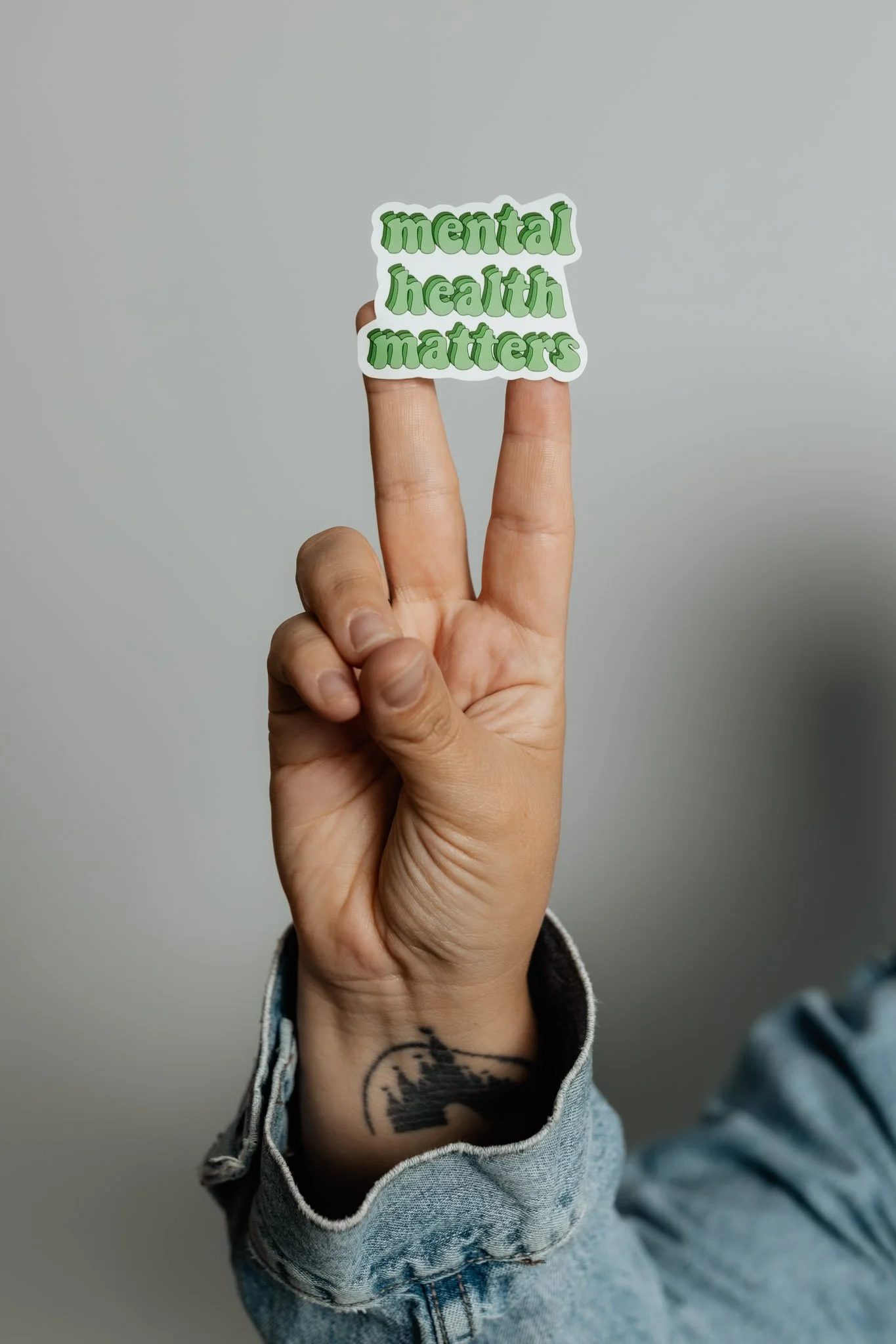 Hand holding a sticker with the words 'mental health matters' written in green, with a tattoo visible on the wrist and part of a denim jacket sleeve.