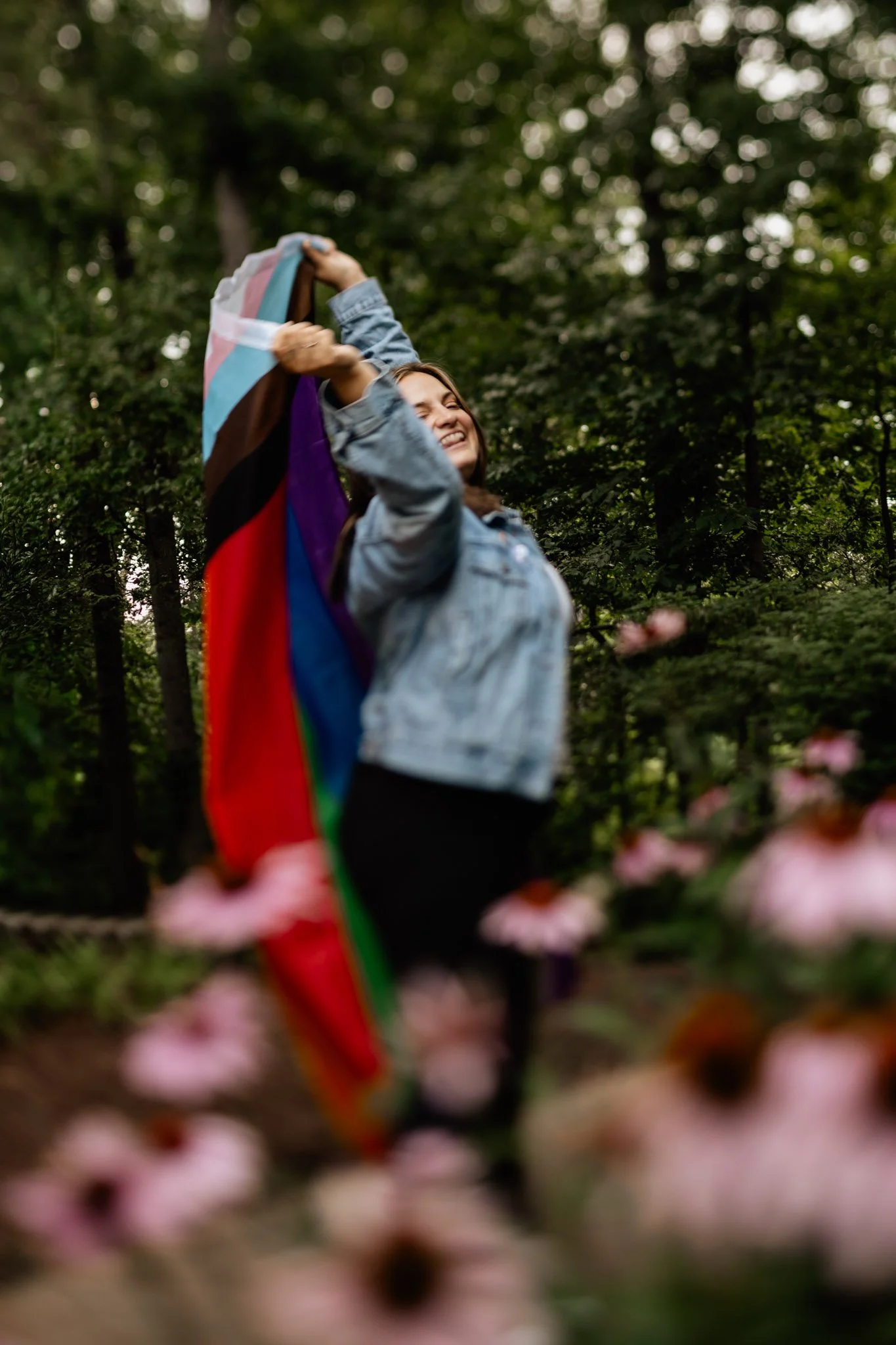 A woman smiling and holding a transgender pride flag outdoors amidst trees, with pink flowers in the foreground.