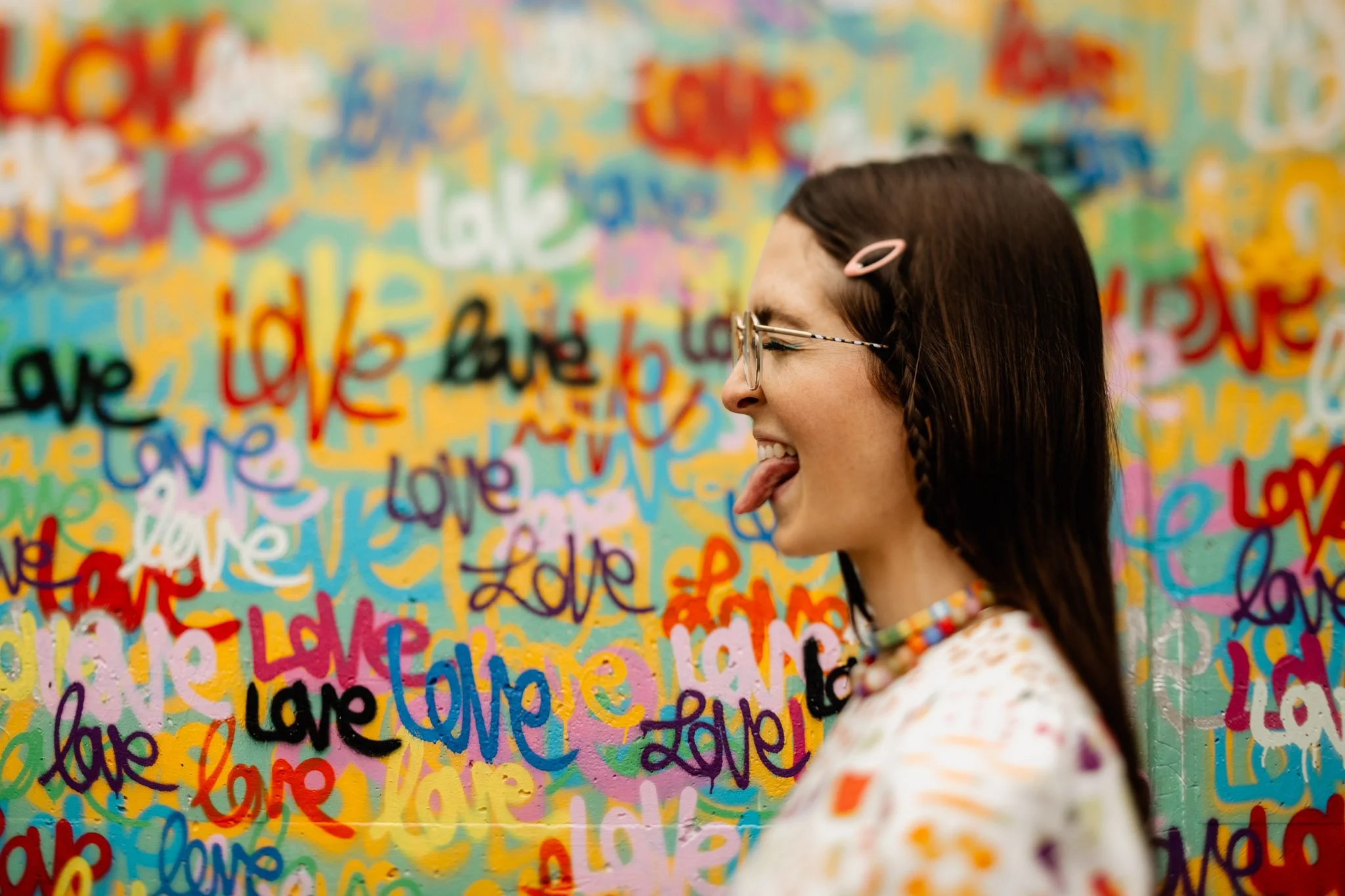 A woman with glasses and a pink hair clip sticking out her tongue, standing in front of a colorful wall with handwritten words like 'love' and 'hope' in various colors.