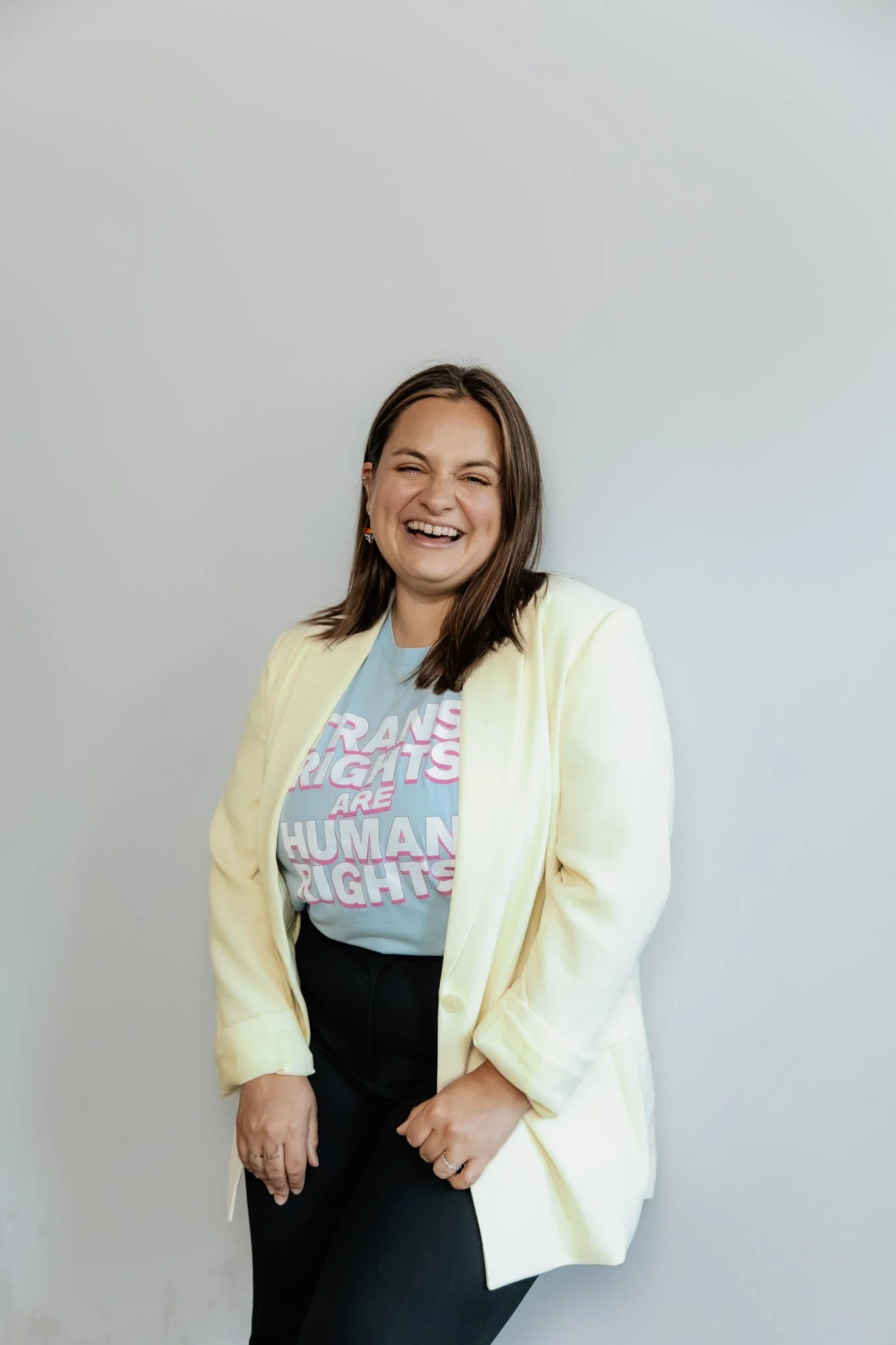A woman standing against a plain light gray wall, laughing with her eyes closed. She has shoulder-length brown hair and is wearing a light yellow blazer over a T-shirt that says "Trans Rights Are Human Rights" in pink and white letters. She is also w