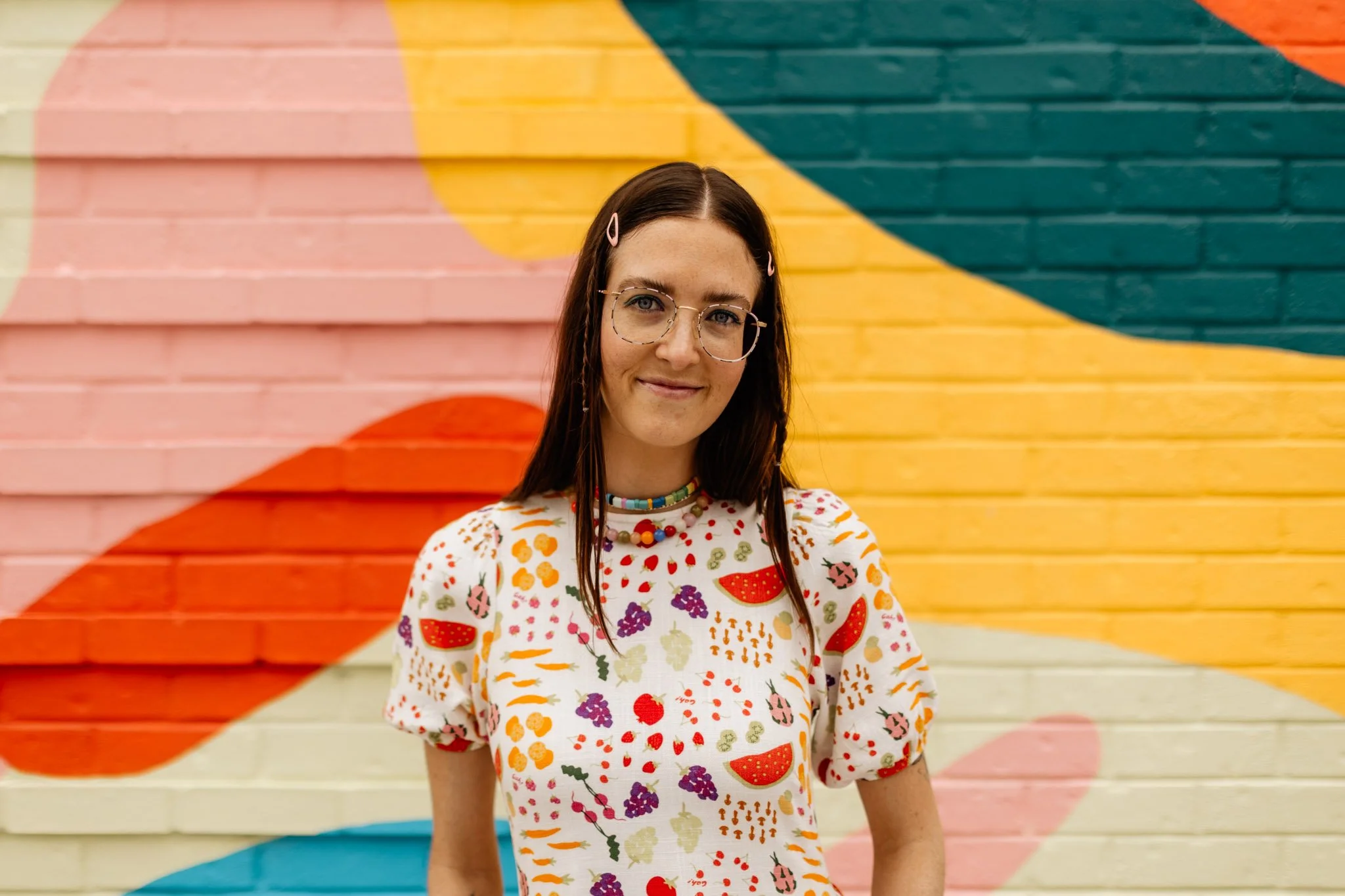 A young woman with long dark hair, wearing glasses, a colorful beaded necklace, and a white dress with a fruit pattern, standing in front of a multicolored graffiti wall.