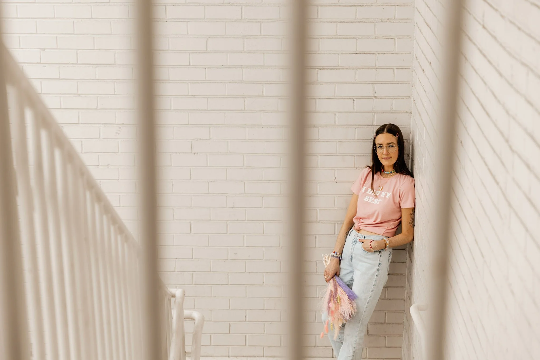 A young woman with long dark hair, wearing glasses, a pink T-shirt, and light blue jeans, leaning against a white brick wall in a stairwell, holding a colorful blanket.