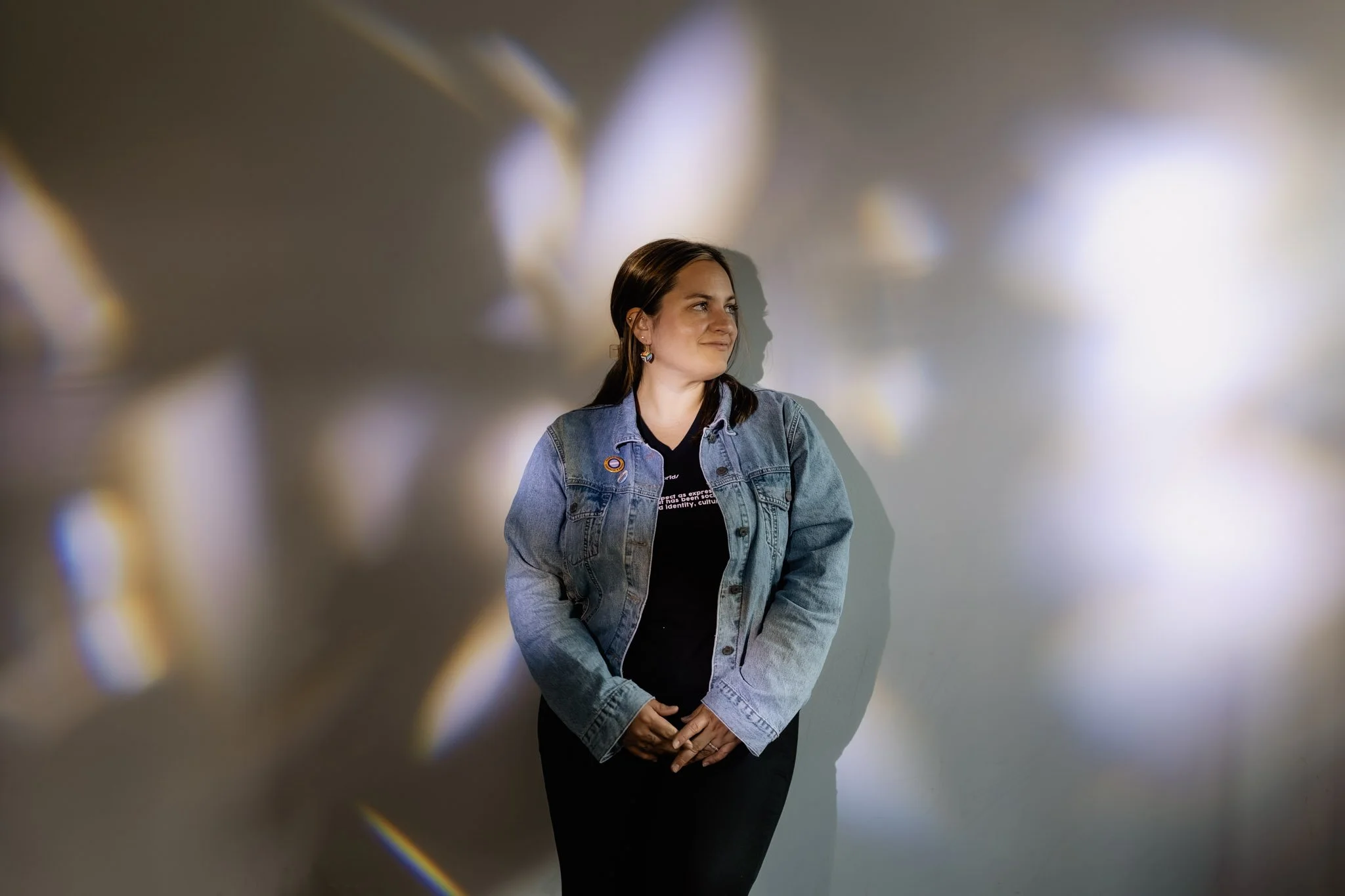 A woman with dark hair, wearing a blue denim jacket and black shirt, stands against a light-colored wall with a colorful bokeh effect. She is smiling slightly and looking to her right.