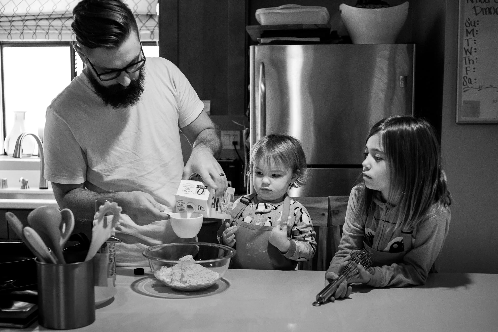 Dad measuring milk into pancake mix with 2 daughters in kitchen