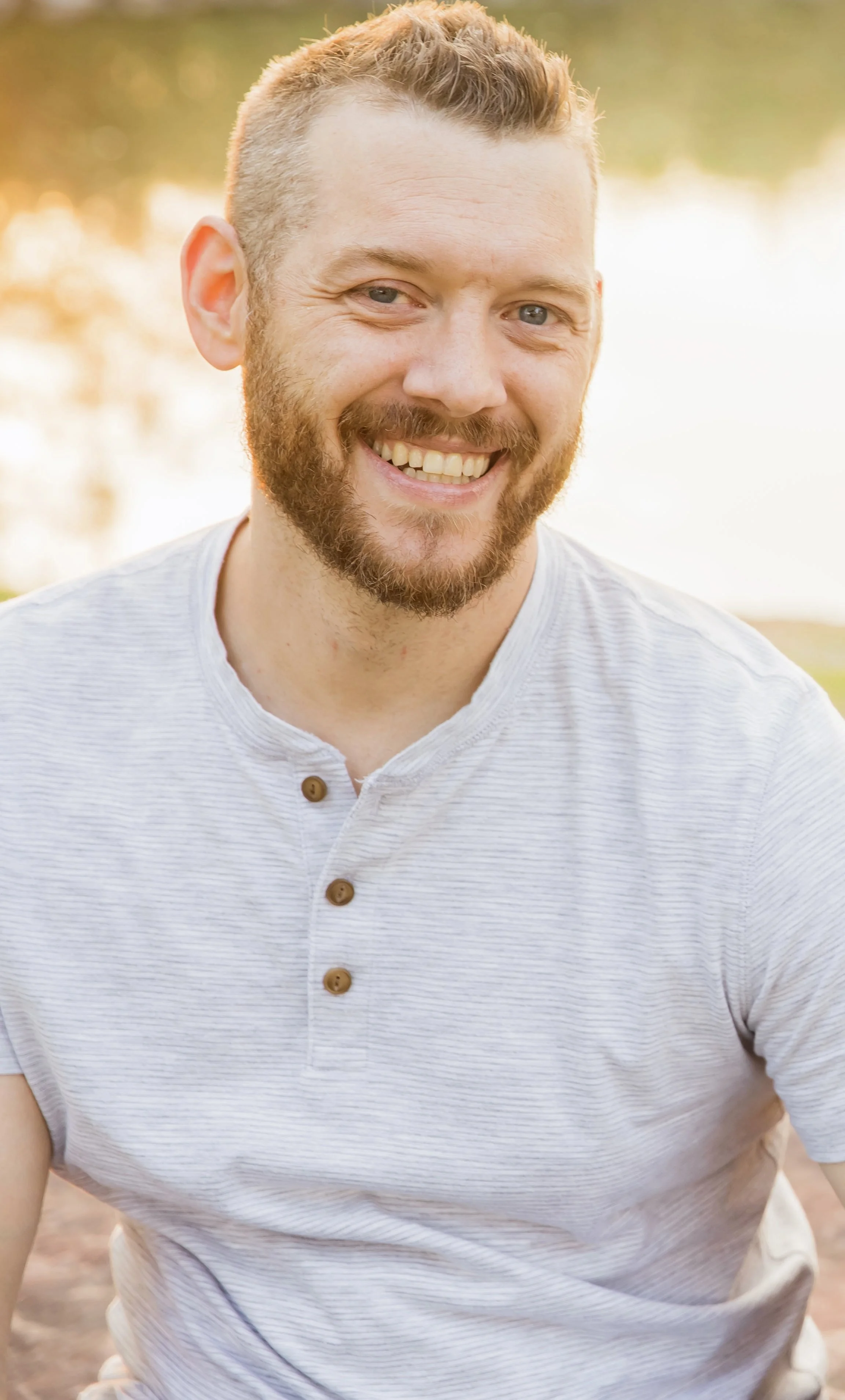 Portrait of a smiling man with a beard and short hair, wearing a white short-sleeve shirt with a button collar, outdoors near a body of water with trees in the background.