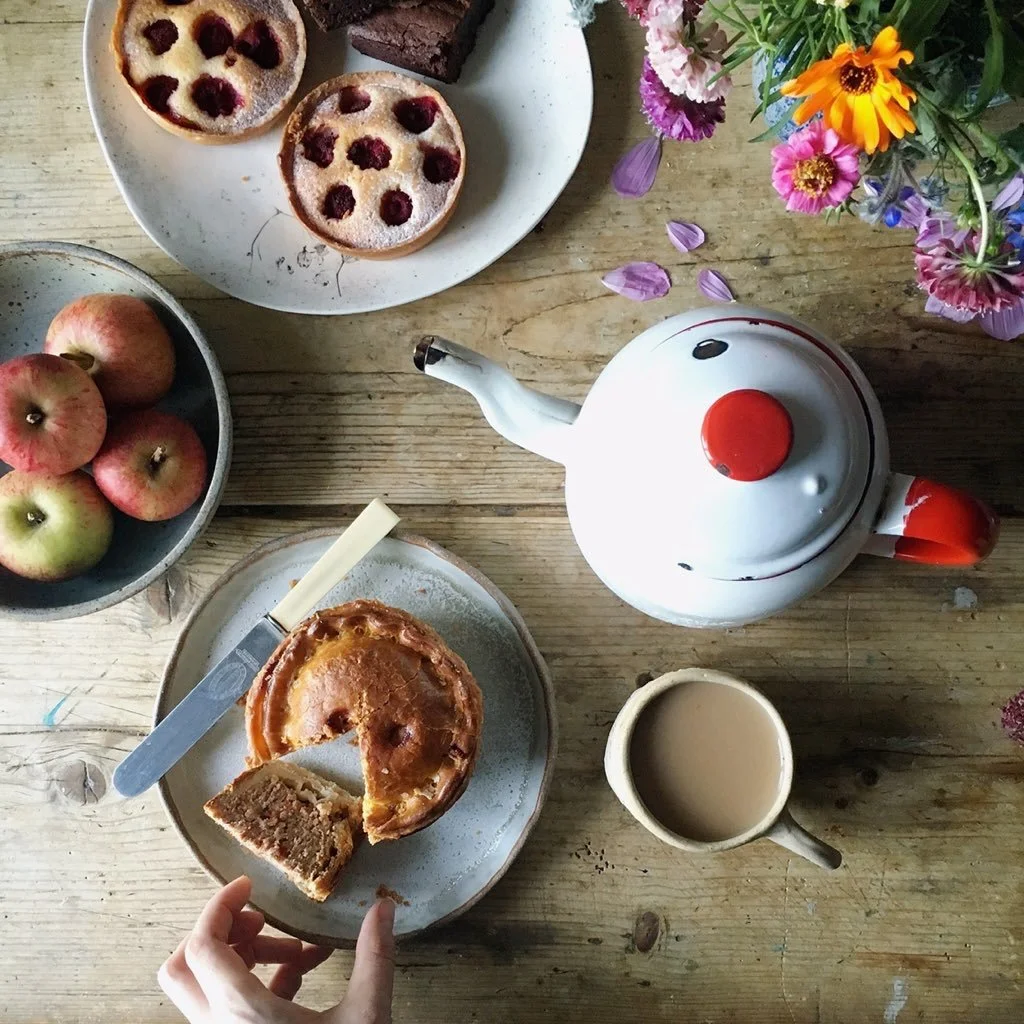 A top-down view of a afternoon tea table with a teapot, a cup of coffee or tea, a sliced cake on a plate, a bowl of three apples, a plate with two cherry-topped pies and a Brays Cottage Norfolk Pork Pie , and a vase of colourful flowers.