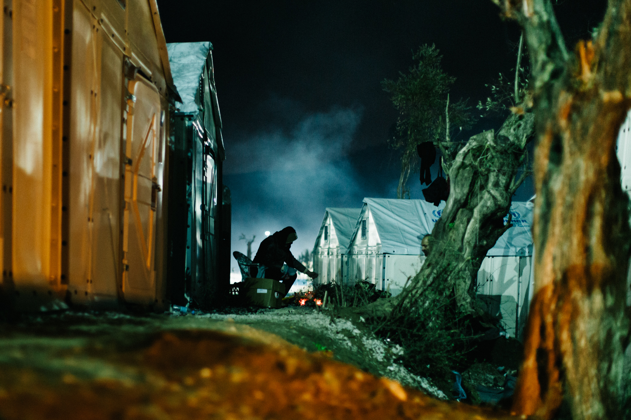Late at night, a man stokes a few embers outside his the shelter his family was given at Moria.