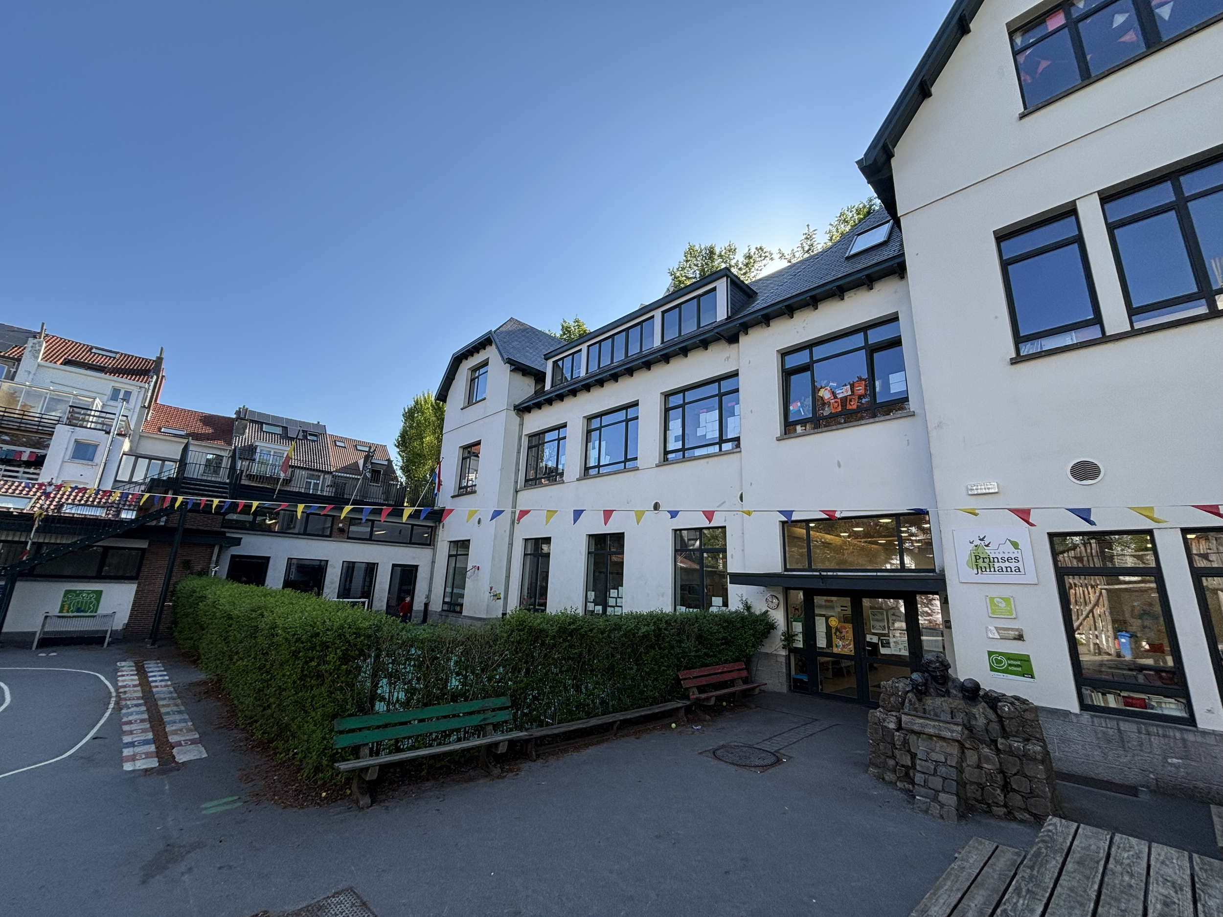 Une école appelée "Princes Juliana" avec des fenêtres modernes, des bancs, un espace de jeux en extérieur avec des drapeaux colorés, et le bâtiment de l'école en arrière-plan.