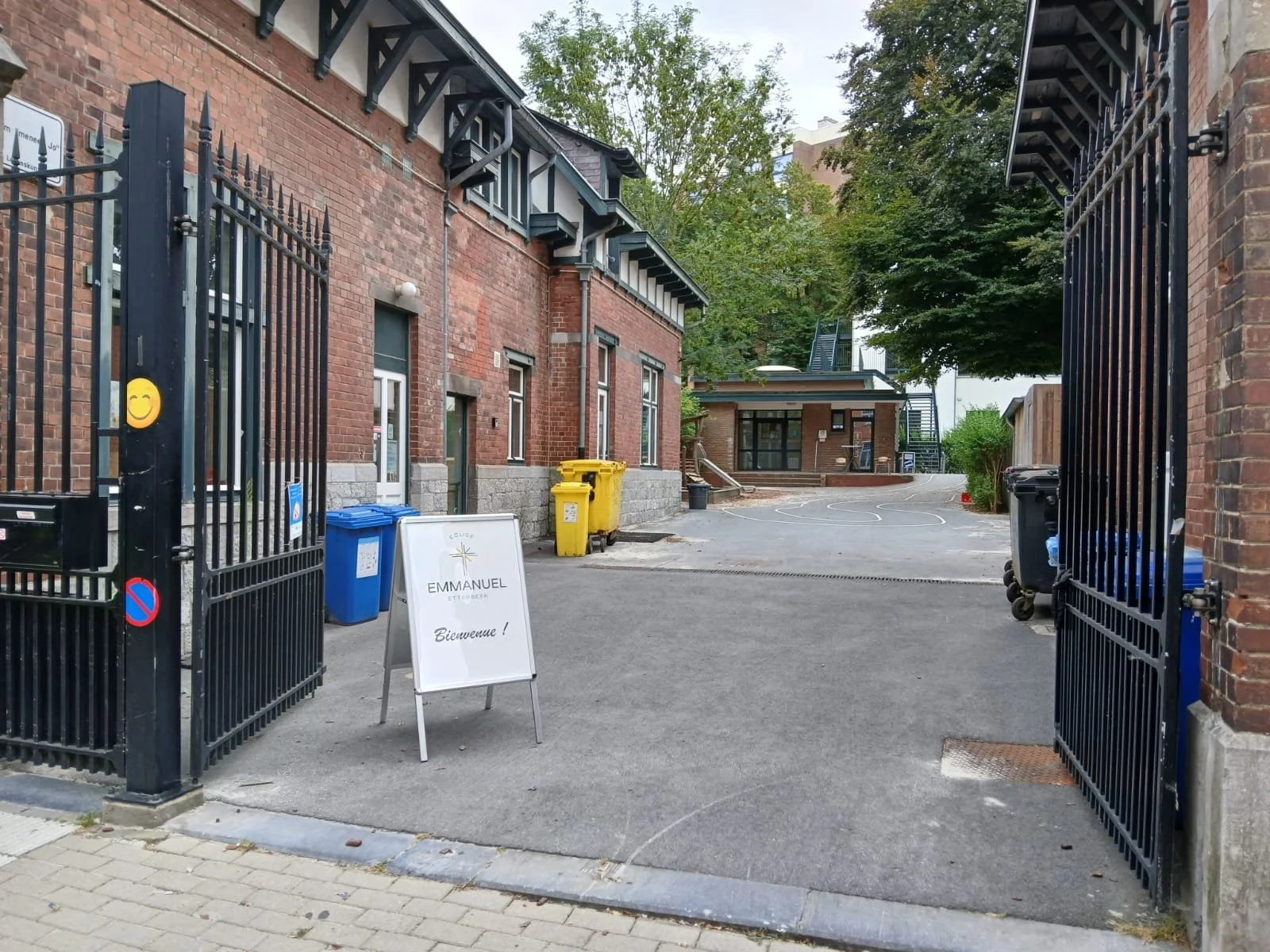Portail noir ouvert donnant accès à une cour avec des bâtiments en briques rouges, des poubelles jaunes et bleues, un panneau blanc avec l'inscription 'Eglise Emmanuel Etterbeek' et 'Bienvenue!'