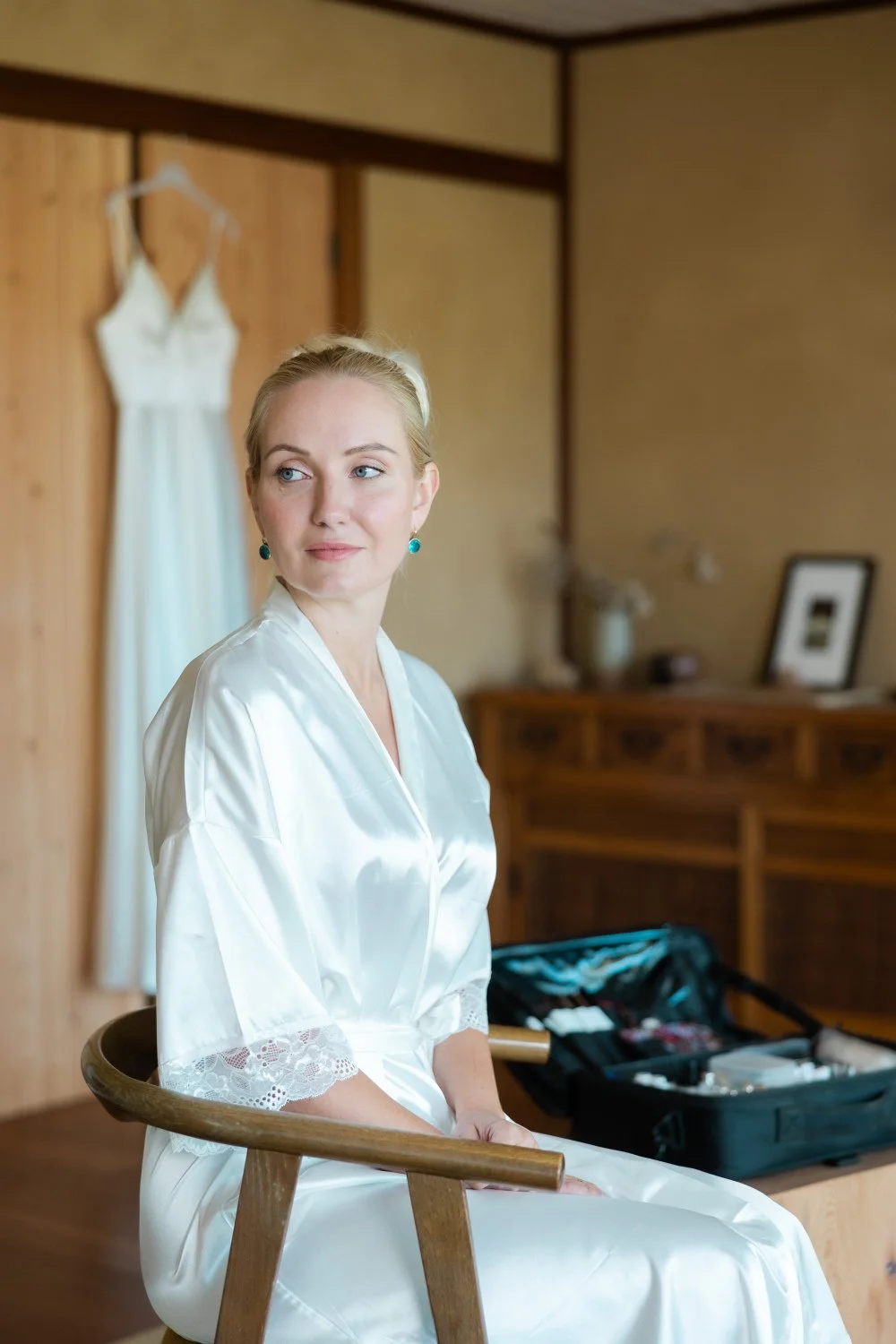 Bride in a white silk robe and natural, glowing skin sits with her dress hanging in the background