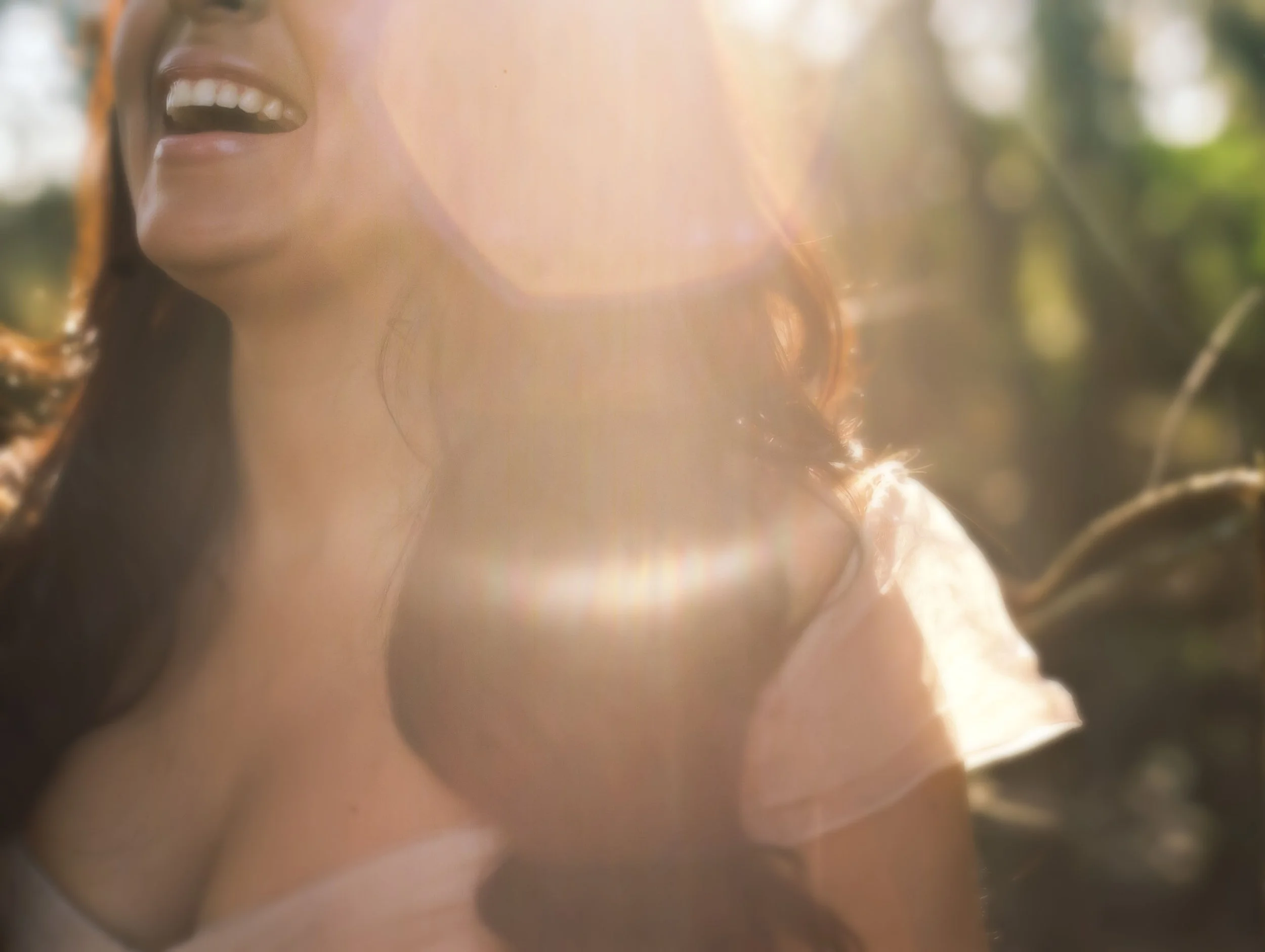 A bride with dark hair in soft waves laughs while sun flares over her shoulder