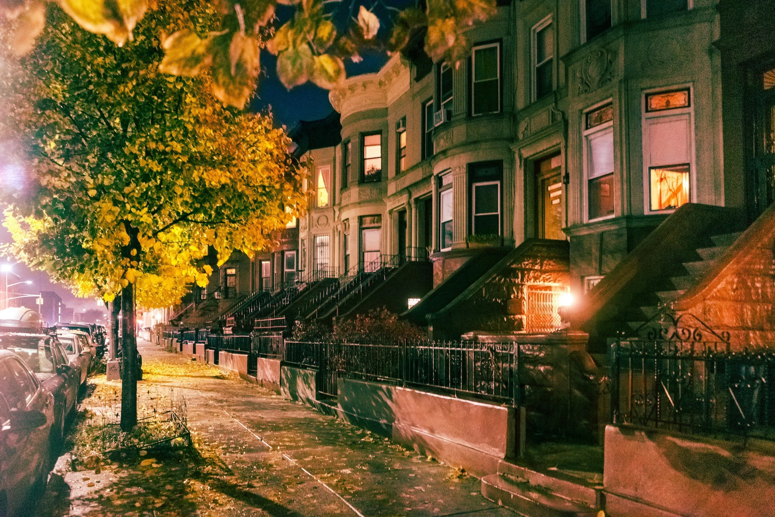 Nighttime view of a residential street with historic row houses, some windows lit, and a tree with yellow leaves illuminated by streetlights. Parked cars line the curb.