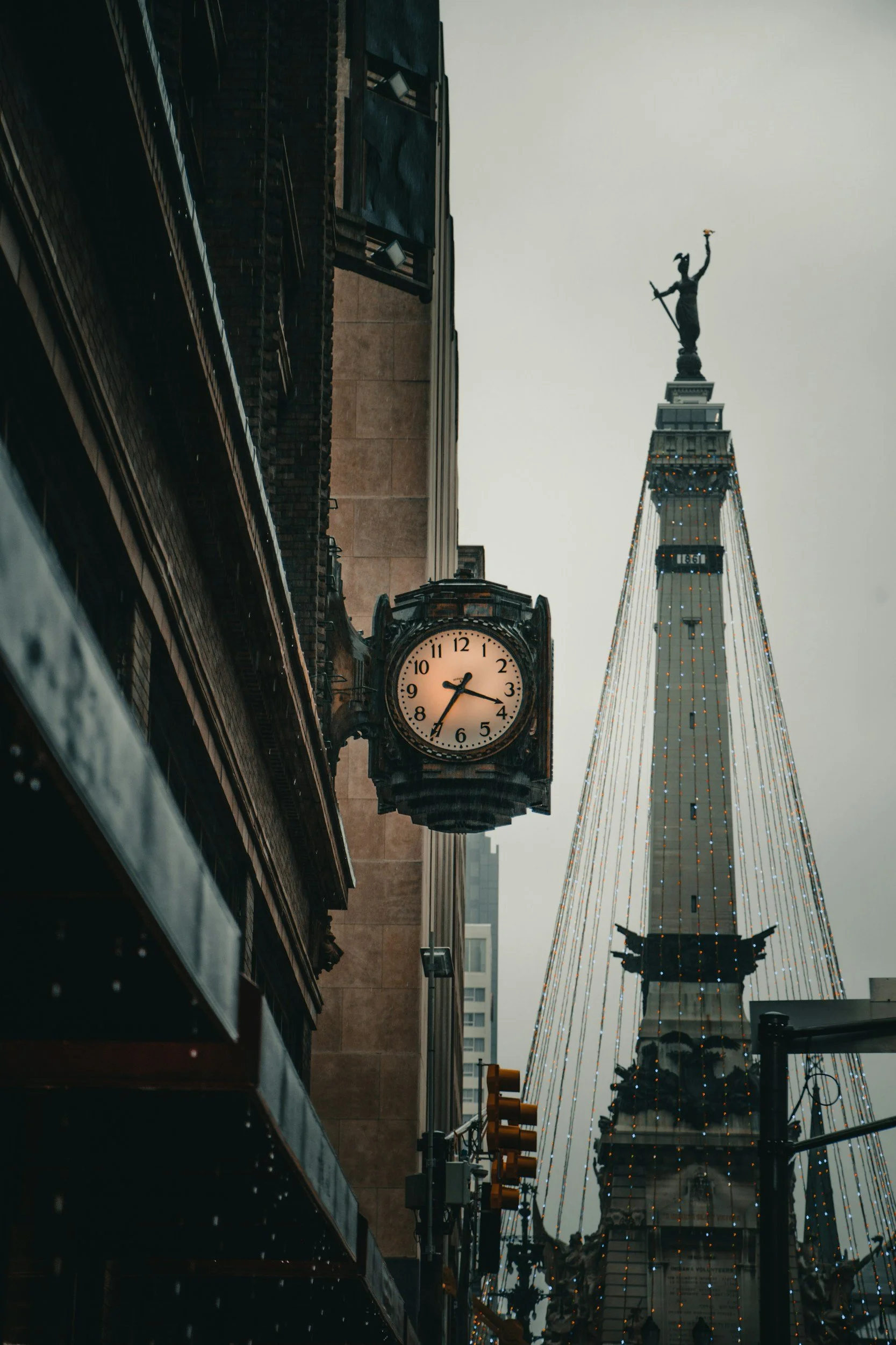 View of a city street featuring a street clock and a tall tower with a statue on top, adorned with string lights, under a cloudy sky.
