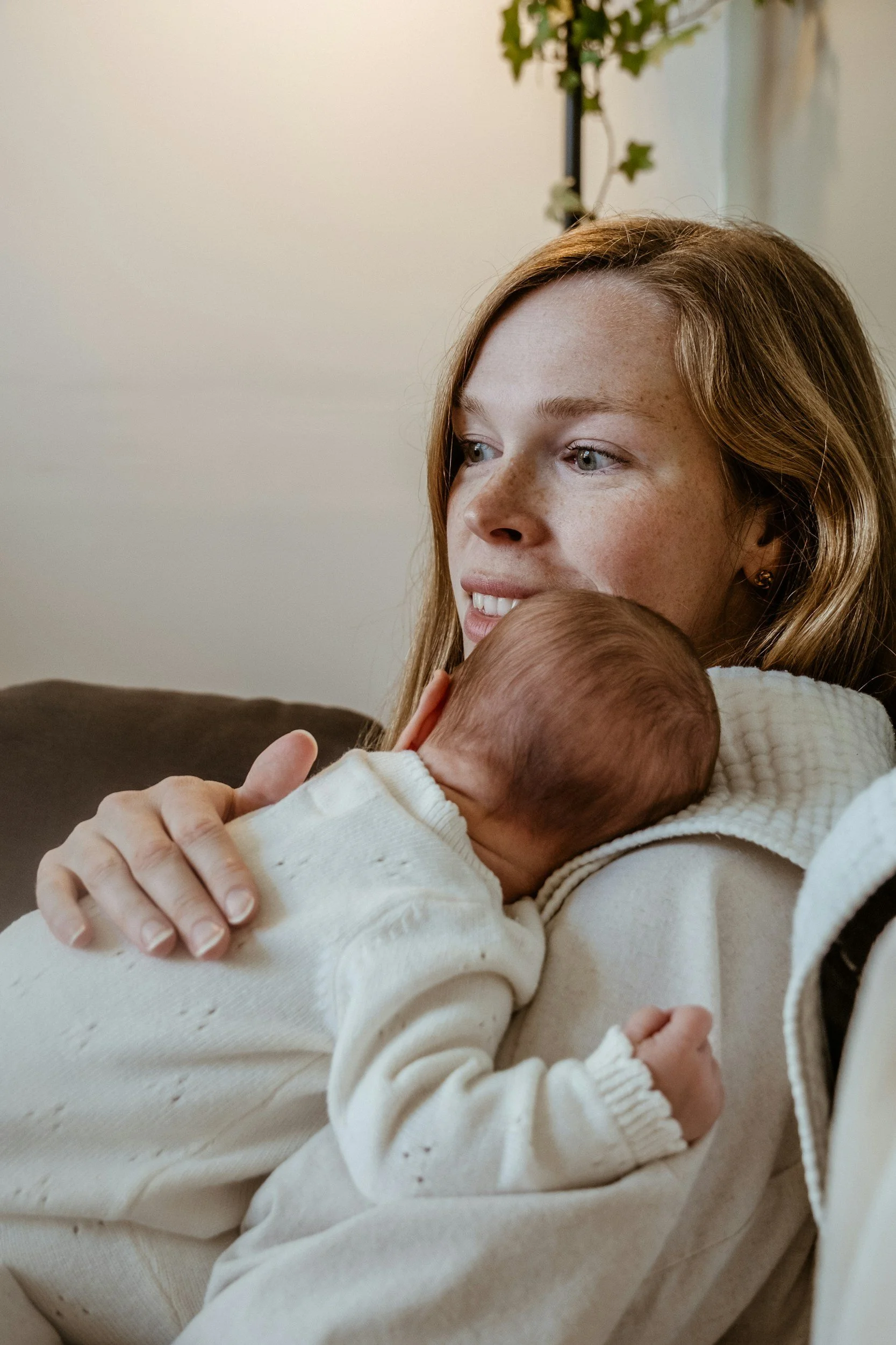 A woman holding a baby close, both dressed in white, sitting on a couch indoors, with a neutral-colored background and a plant in the background.
