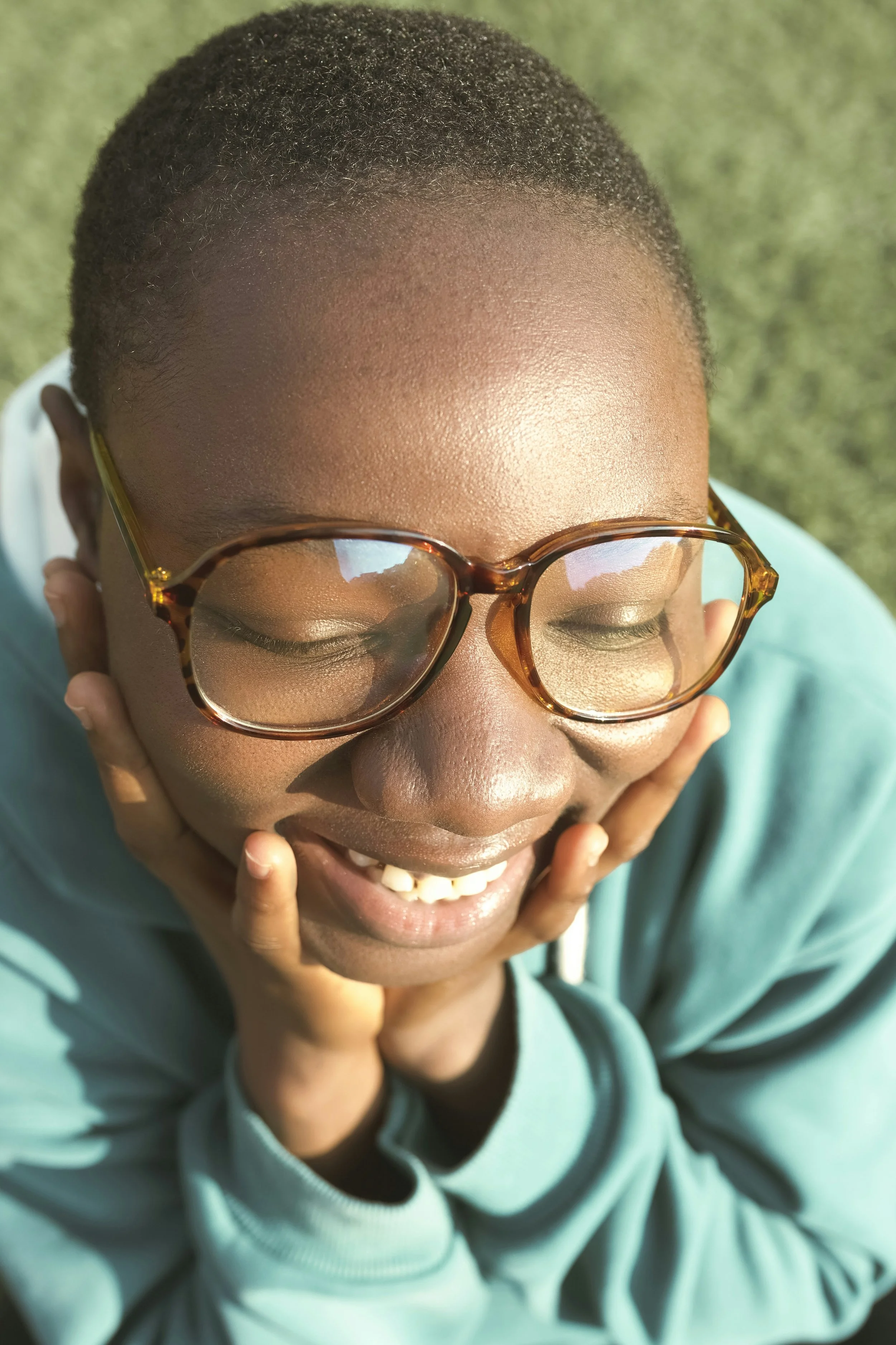 Close-up of a smiling person with glasses, resting their chin on their hands, outside on sunny day.
