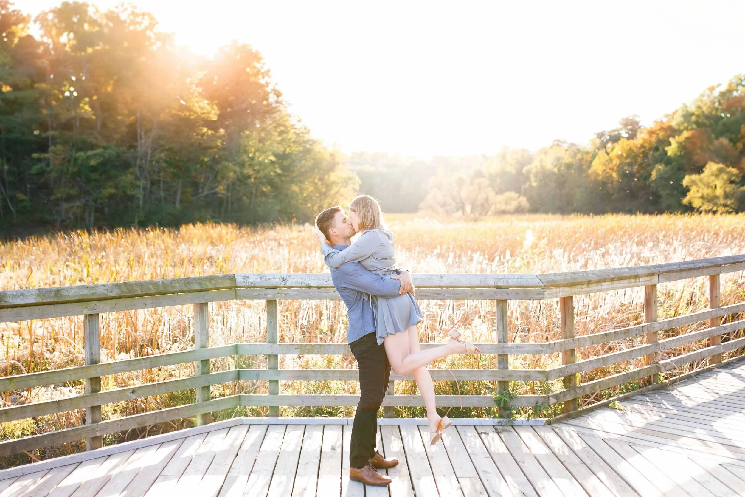 Couple sharing a kiss while he lifts her on the boardwalk at Grindstone Marsh Trail during their Burlington engagement session