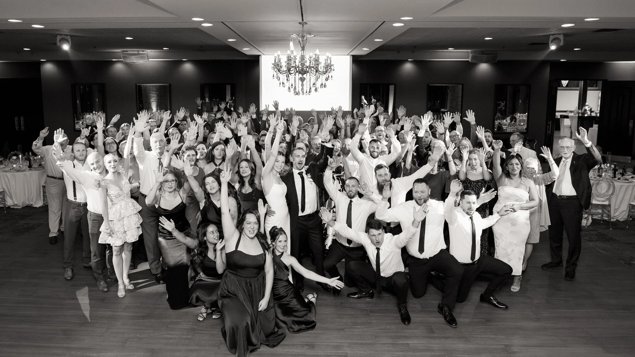Black and white reception crowd portrait beneath chandelier on the dance floor at The Manor Event Venue wedding in King, Ontario