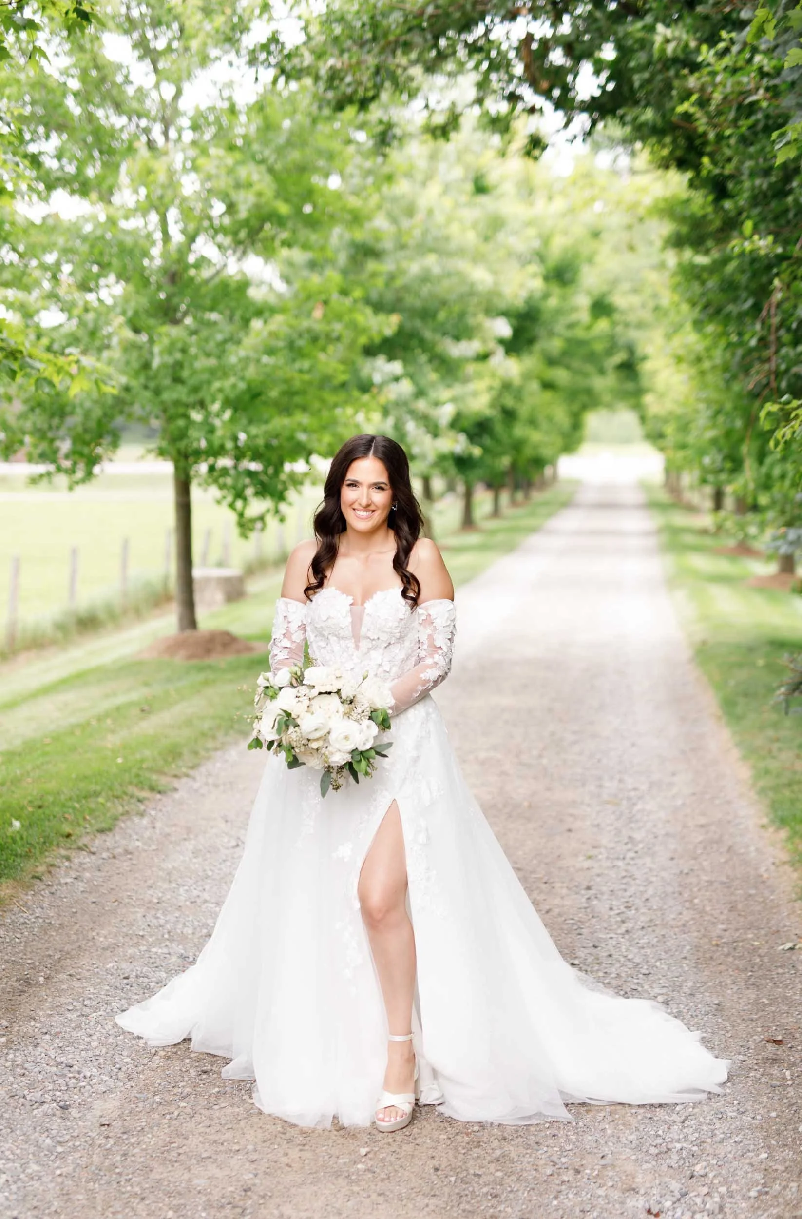Bride standing on the tree-lined driveway holding her bouquet at a Cambium Farms wedding in Alton, Ontario