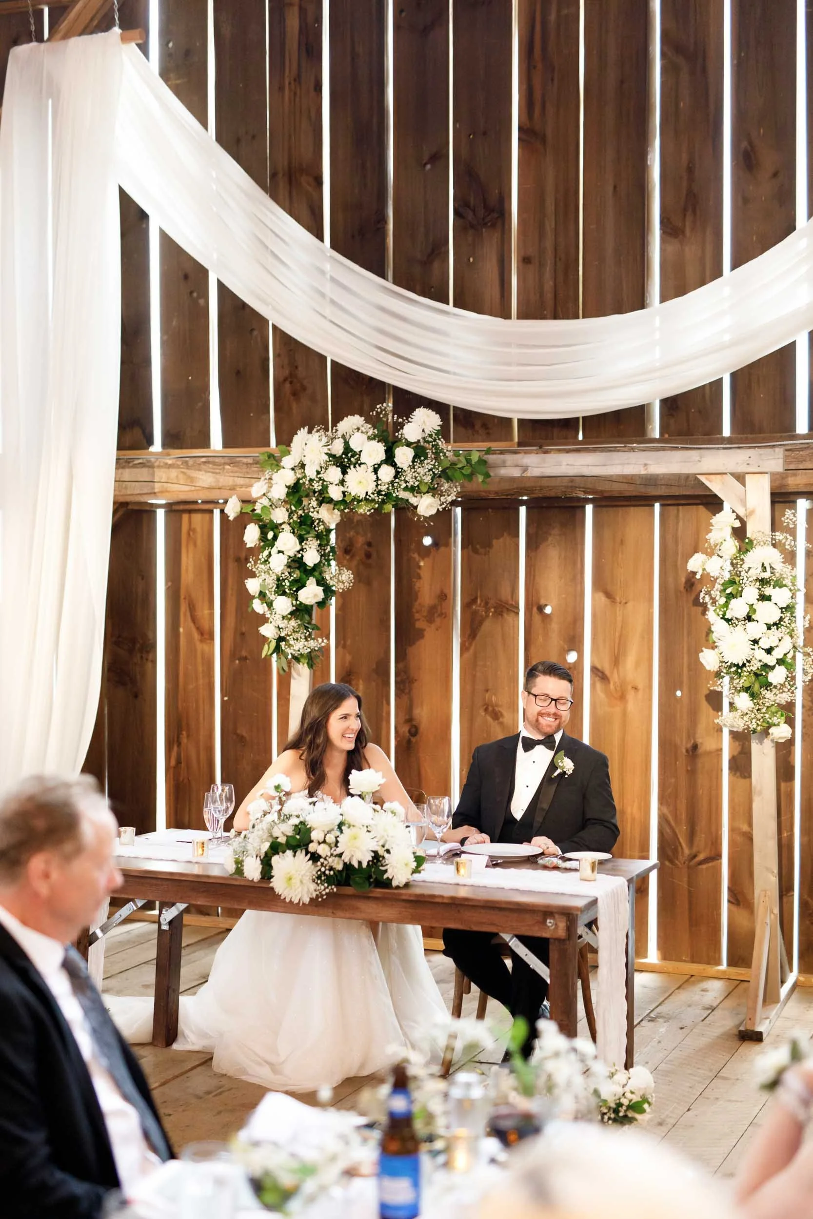 Bride and groom seated at the sweetheart table during speeches at a Cambium Farms wedding in Alton, Ontario