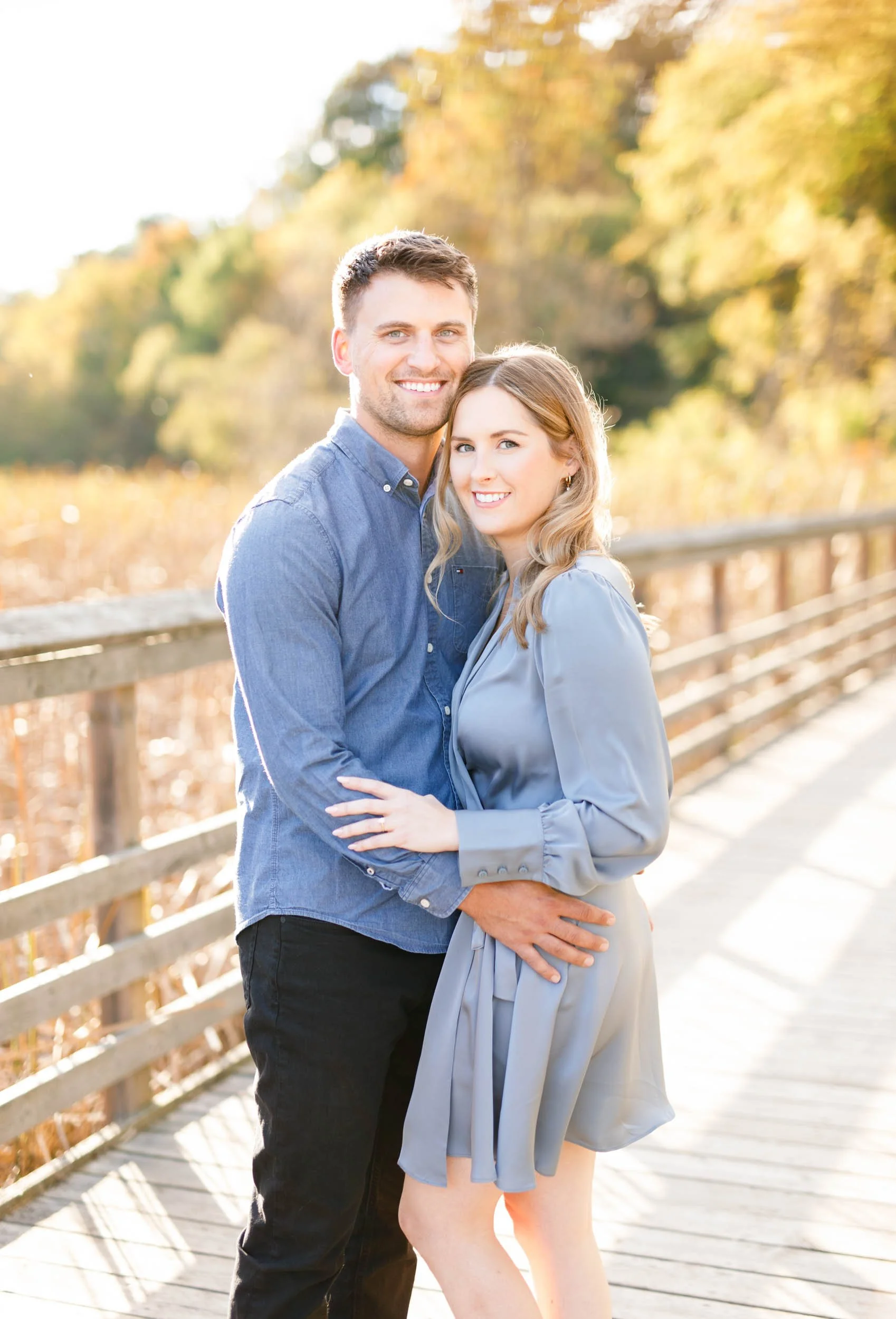 Smiling engaged couple embracing on the boardwalk at Grindstone Marsh Trail in Burlington Ontario during a fall engagement shoot