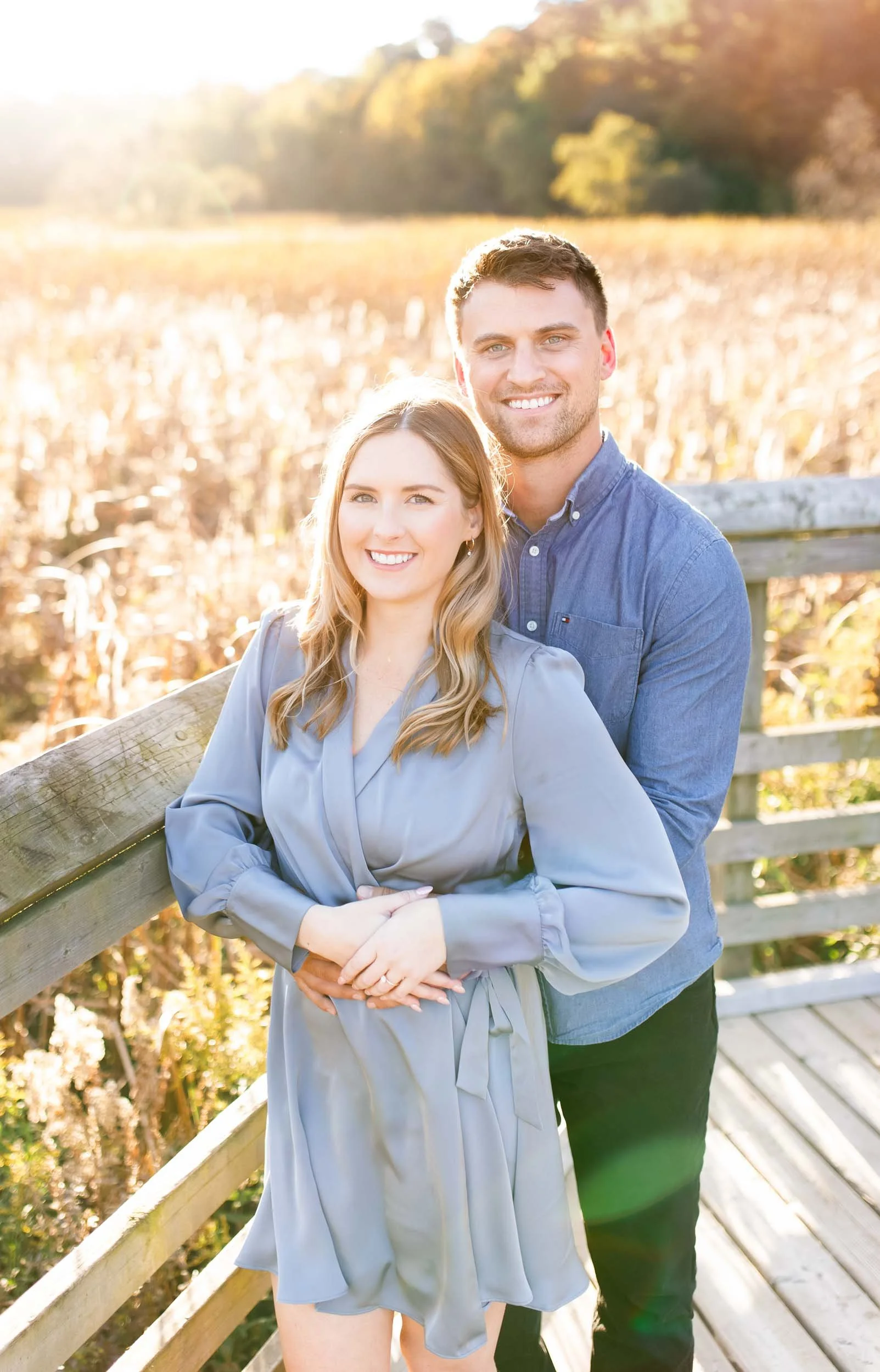 Smiling engaged couple posing together at Grindstone Marsh Trail in Burlington Ontario with golden autumn light