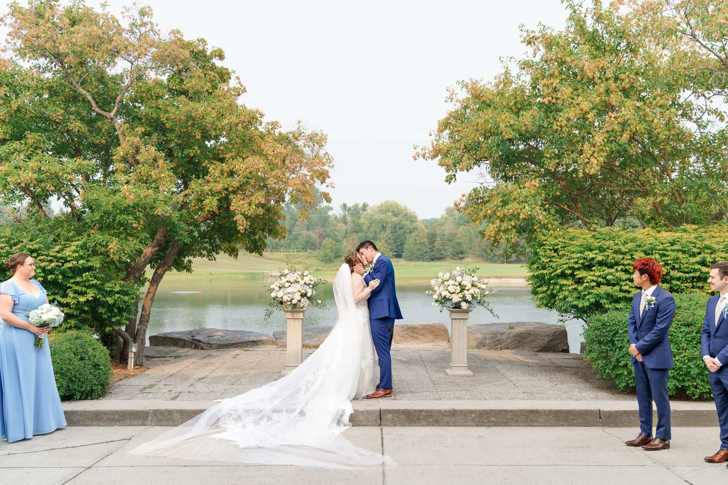 Newlyweds sharing a kiss beside lakeside florals at The Manor Event Venue Wedding ceremony in King, Ontario