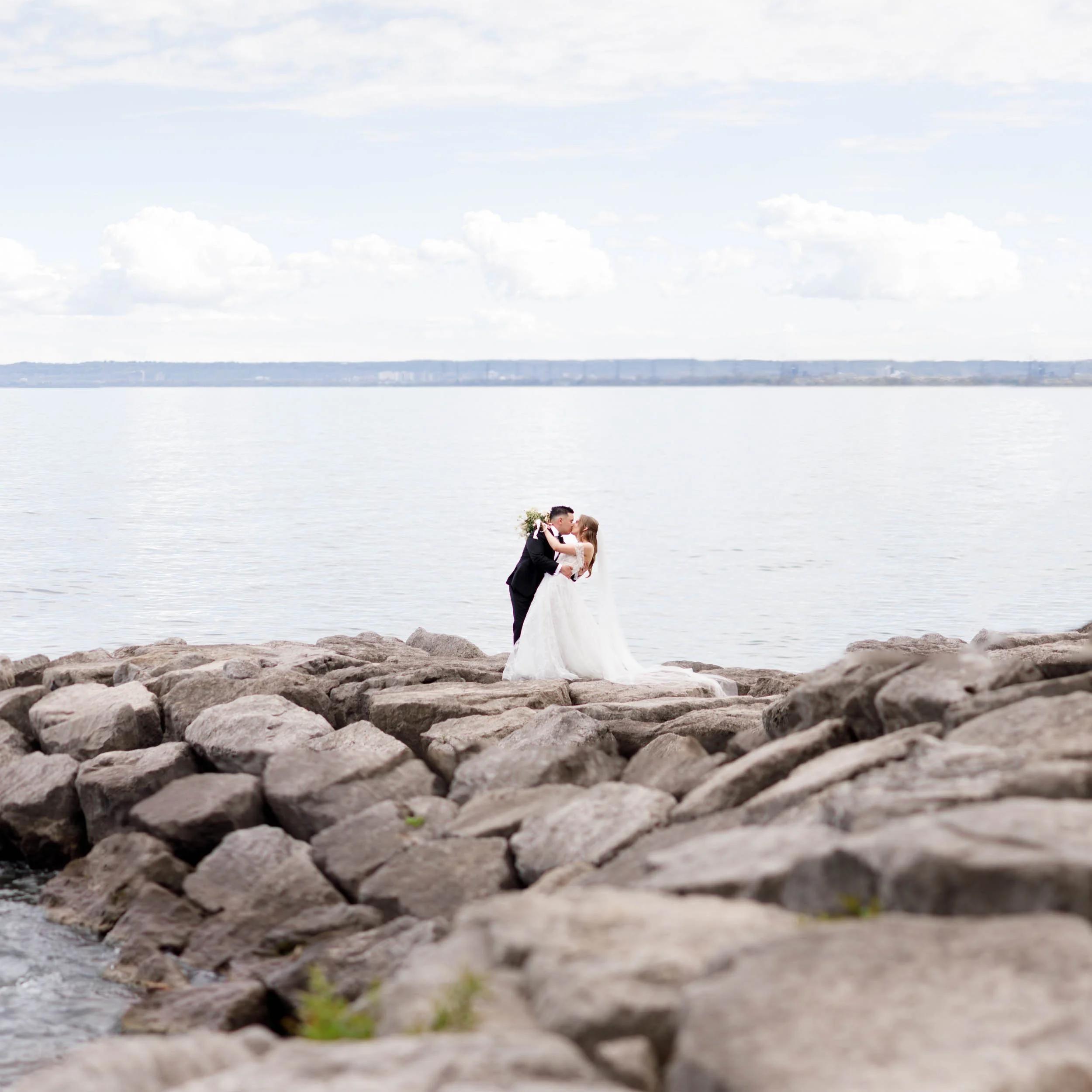 Cinematic wide shot of a bride and groom embracing on the waterfront rocks at a Pearle Hotel wedding in Burlington, Ontario