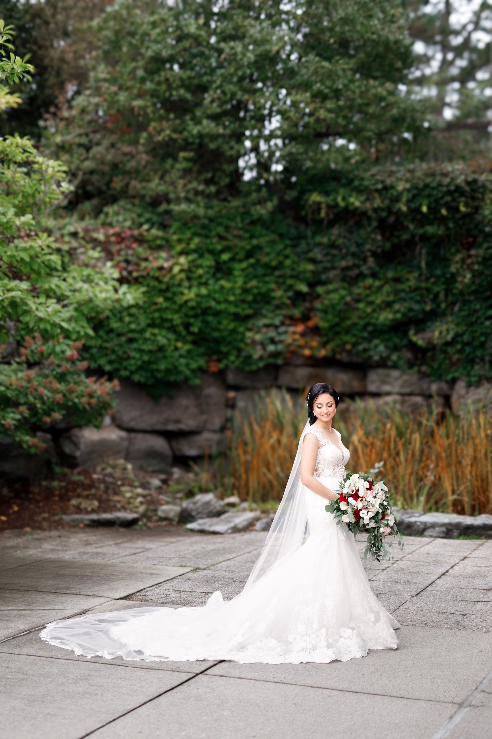 Bride posing with red and white bouquet against stone backdrop at The Manor Event Venue wedding in King, Ontario