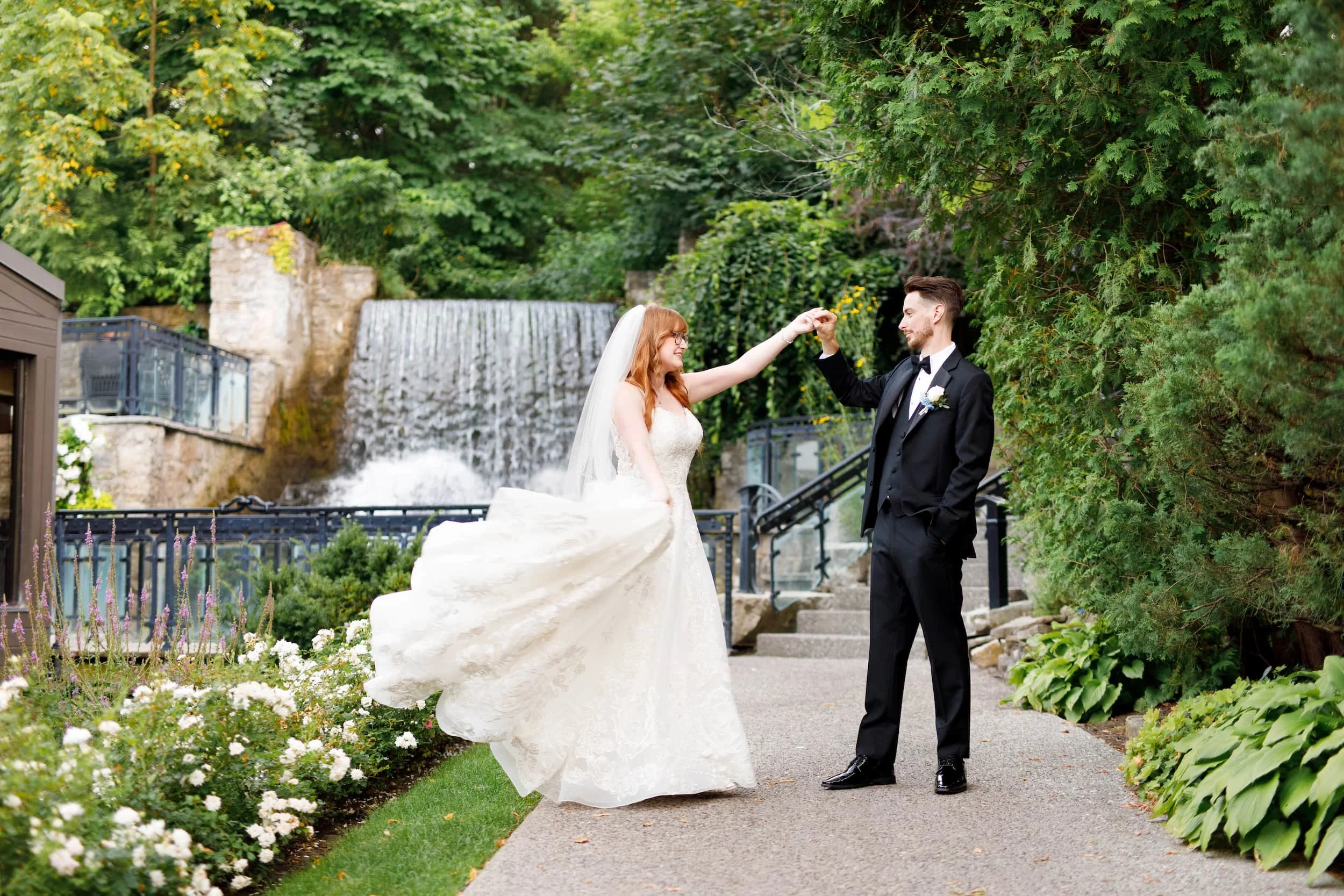 Groom twirling bride with flowing wedding gown beside the waterfall at the Ancaster Mill in Hamilton, Ontario