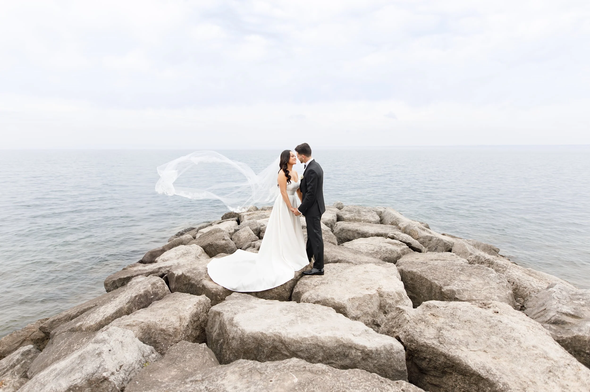 Bride with flowing veil and groom standing on the lakeside rocks at the Pearle Hotel in Burlington