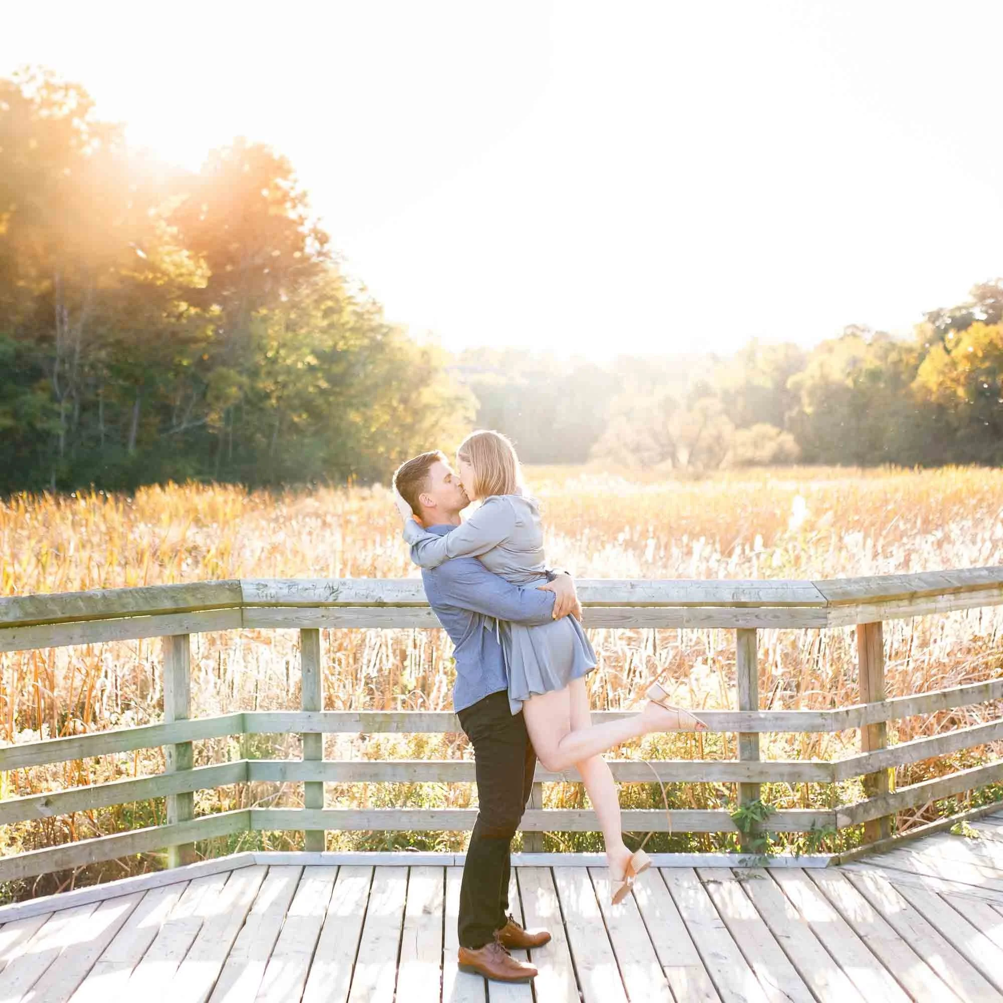 Groom lifting bride during a sunset engagement session on the Grindstone Marsh Trail boardwalk in Burlington - View Engagement Portfolio