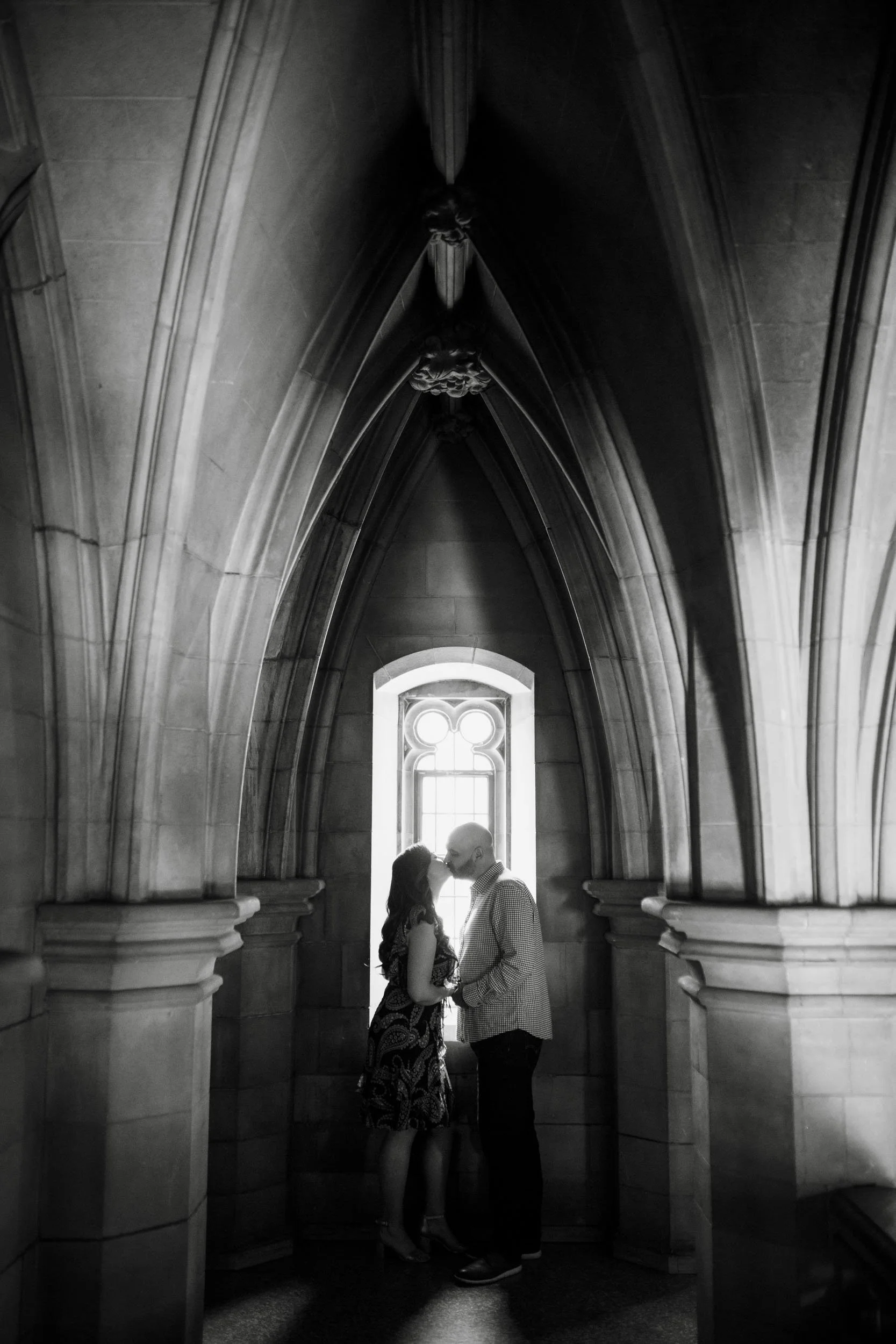 Black and white engagement portrait beneath Gothic arch at Knox College, University of Toronto in Toronto, Ontario