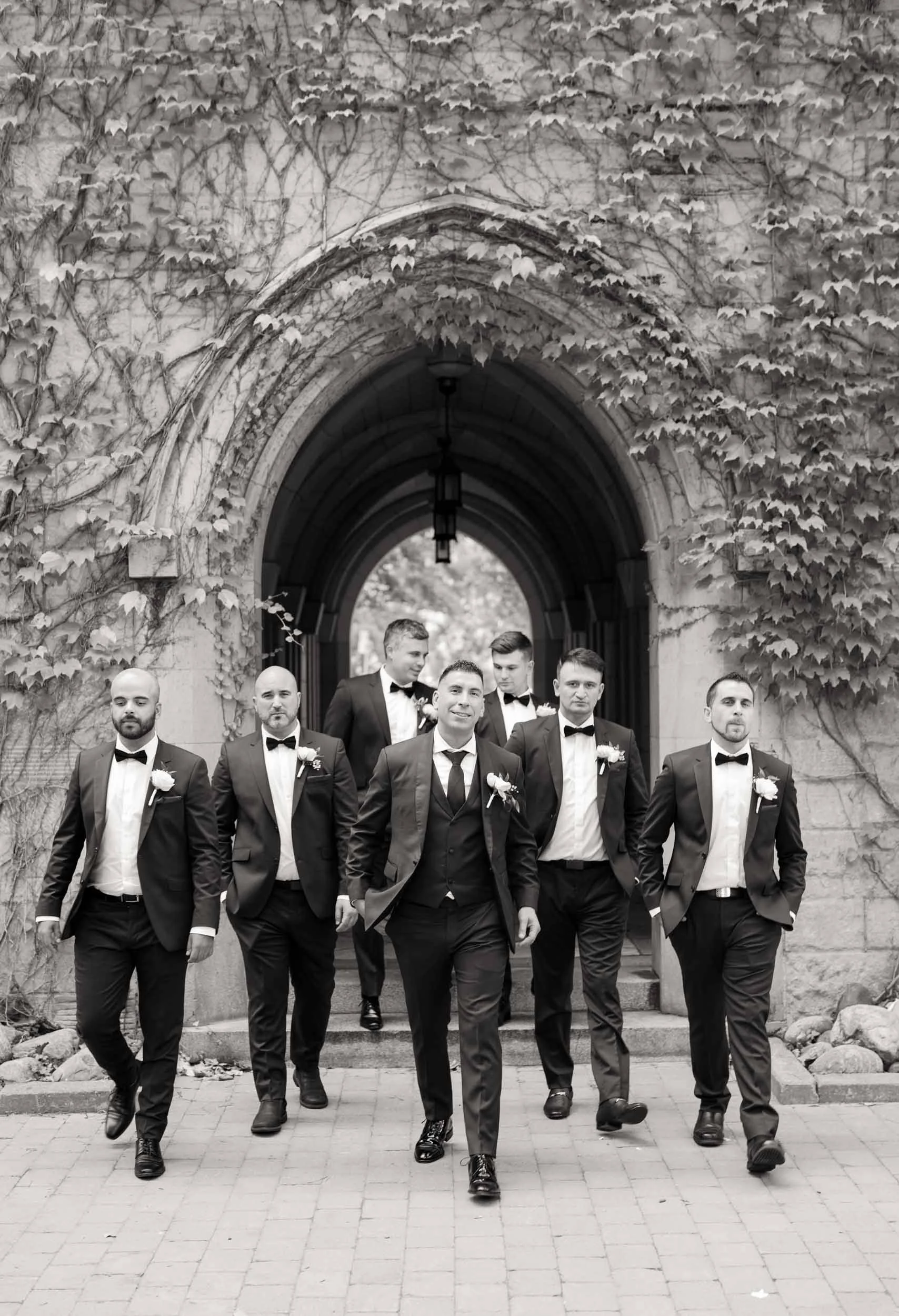 Black and White shot of Groomsmen walking beneath ivy-covered arch at St. Michael’s College, University of Toronto in Toronto, Ontario