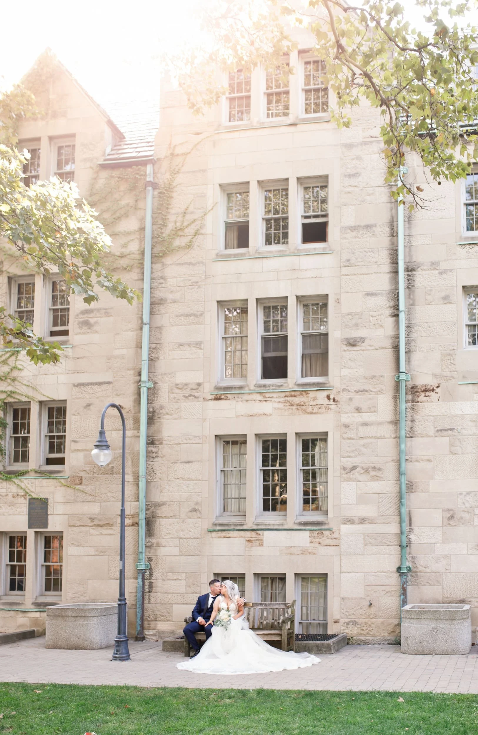 Wide angle of bride and groom sitting under Architectural detail of limestone façade at St. Michael’s College, University of Toronto in Toronto, Ontario